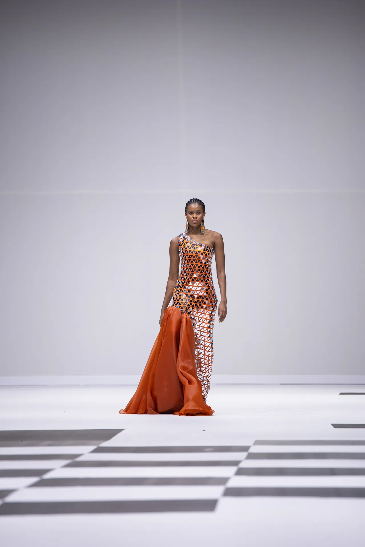 Model walking the runway in an orange geometric-patterned one-shoulder gown, holding a trailing orange fabric piece.