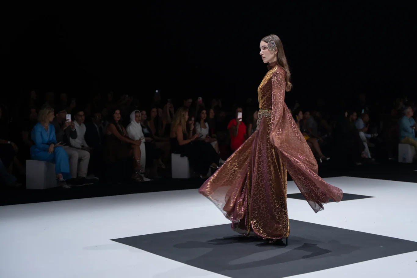 Model in a burgundy-gold embroidered gown walks the runway with a dark audience backdrop.