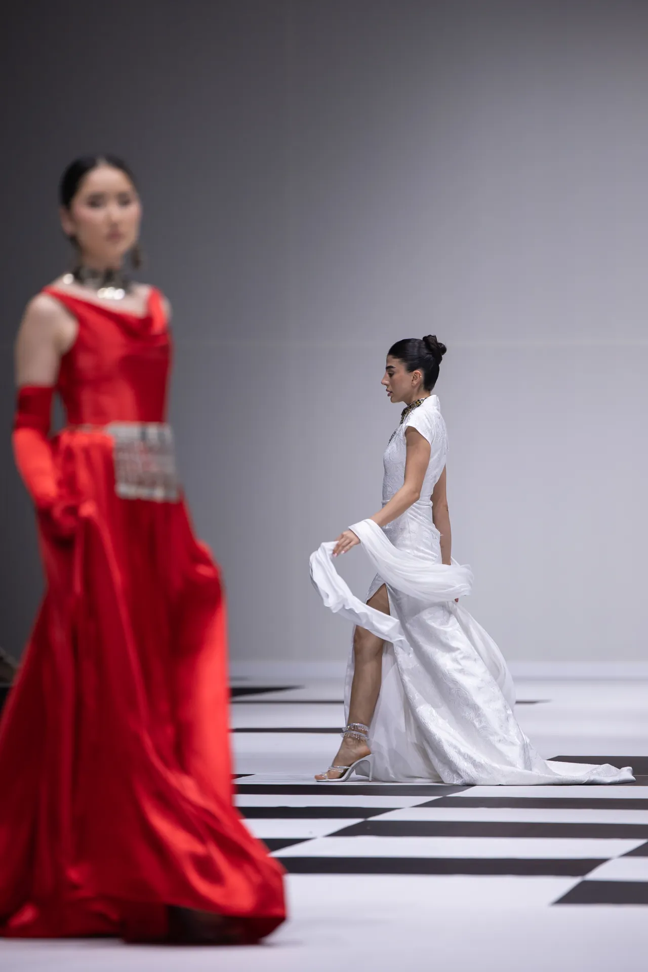 Model in a white gown walking the runway on a black-and-white checkered floor, blurred red-dressed model in foreground.