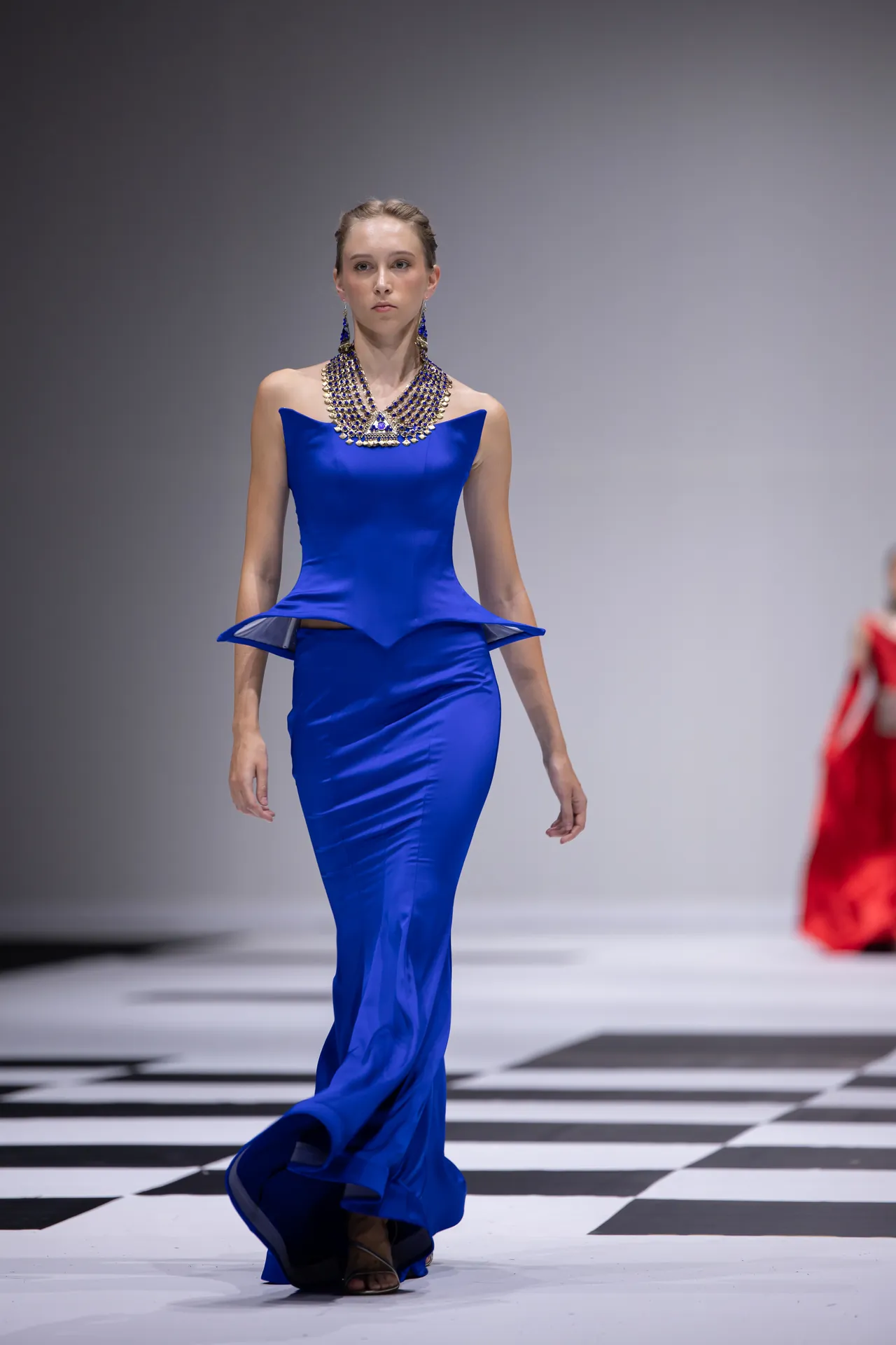 Model in a royal blue gown with a peplum waist and a heavy jeweled necklace walks a black-and-white checkerboard runway.