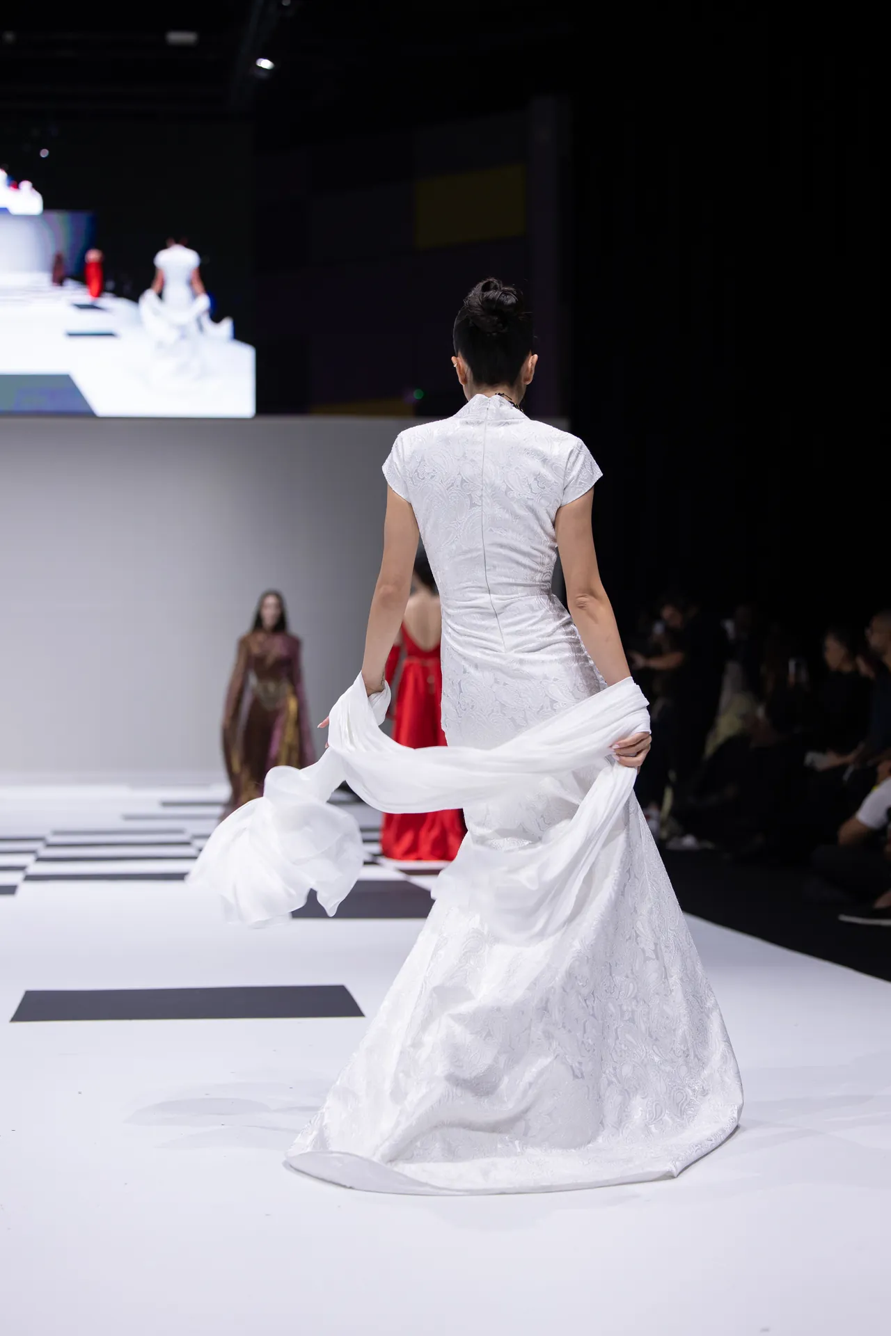 Back view of a model in a white lace gown walking a runway, white fabric trailing as she swirls it behind her.