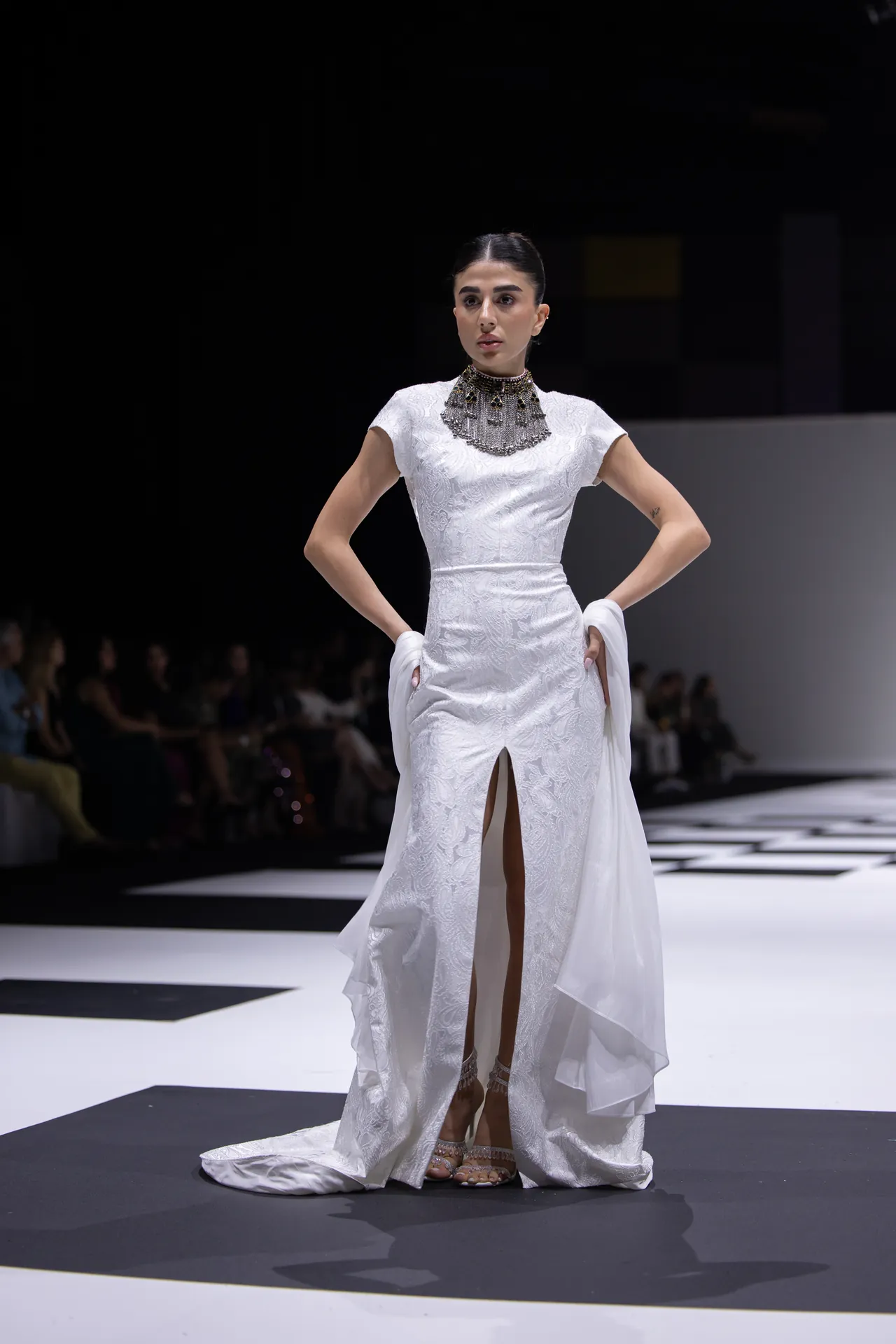 Model on a fashion runway wearing a white lace gown with high slit and long train, paired with a statement necklace and gloves.