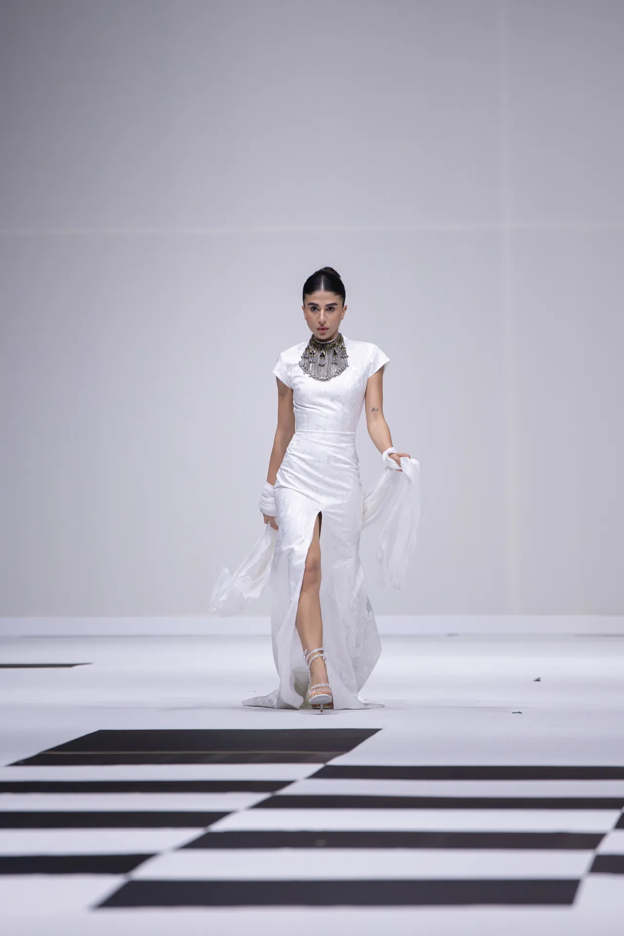 Model in a white, floor-length gown with a high slit and heavy necklace walking a stark white runway with a draped train of fabric nearby.