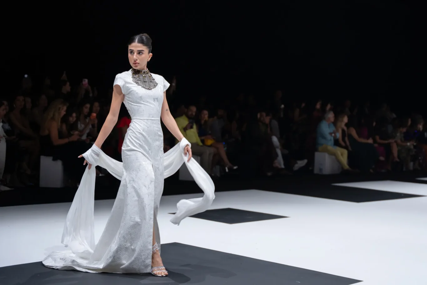 Fashion model in a white, floor-length gown on a runway, cape-like fabric flowing as she walks with a dark audience backdrop.