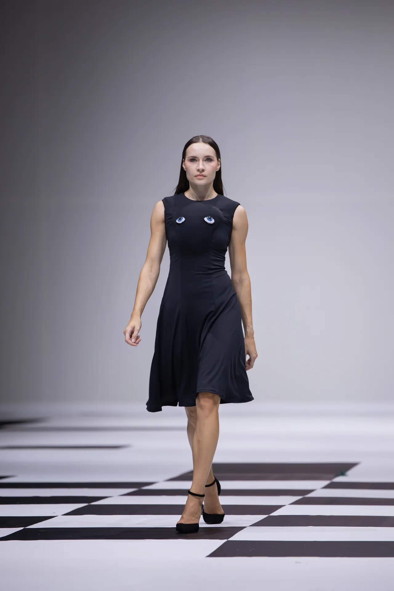 Model walking on a runway in a black sleeveless dress with blue eye designs on the chest, checkerboard floor beneath.