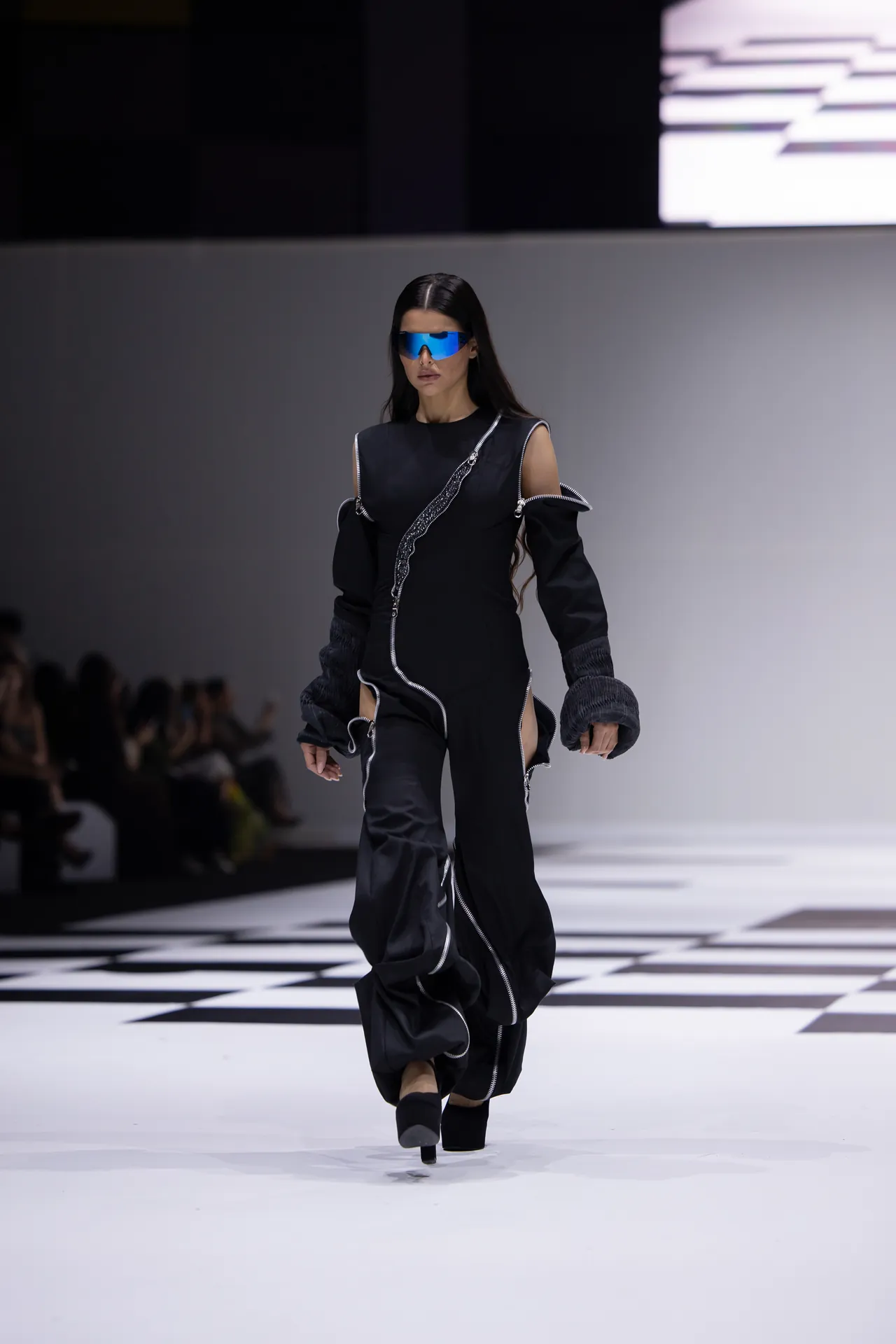 Model walks a black, avant-garde outfit with oversized sleeves and silver zippers on a checkered runway, wearing blue visor sunglasses.
