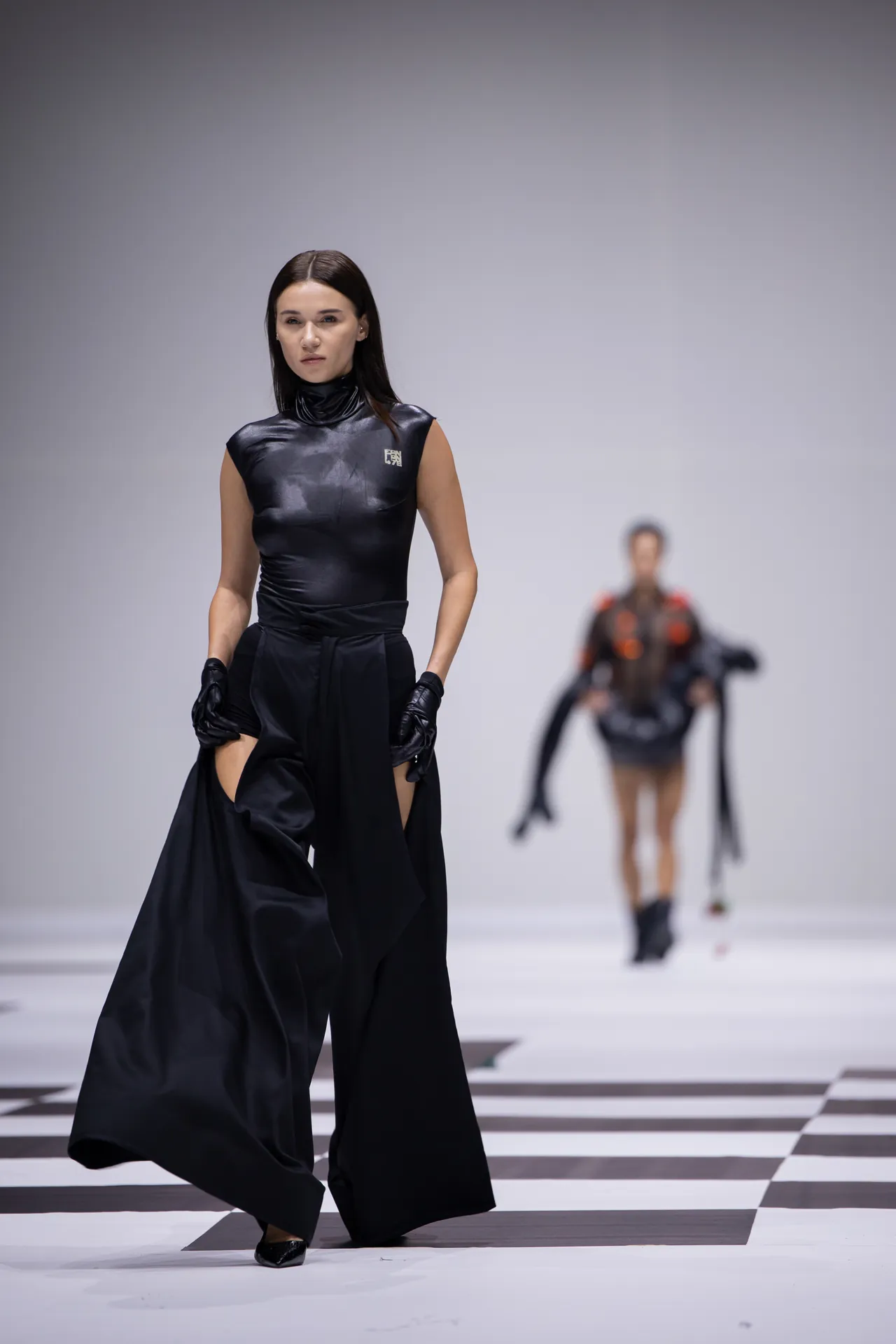 Model walking a black avant-garde dress on a checkered runway, others blurred in background in a fashion show.