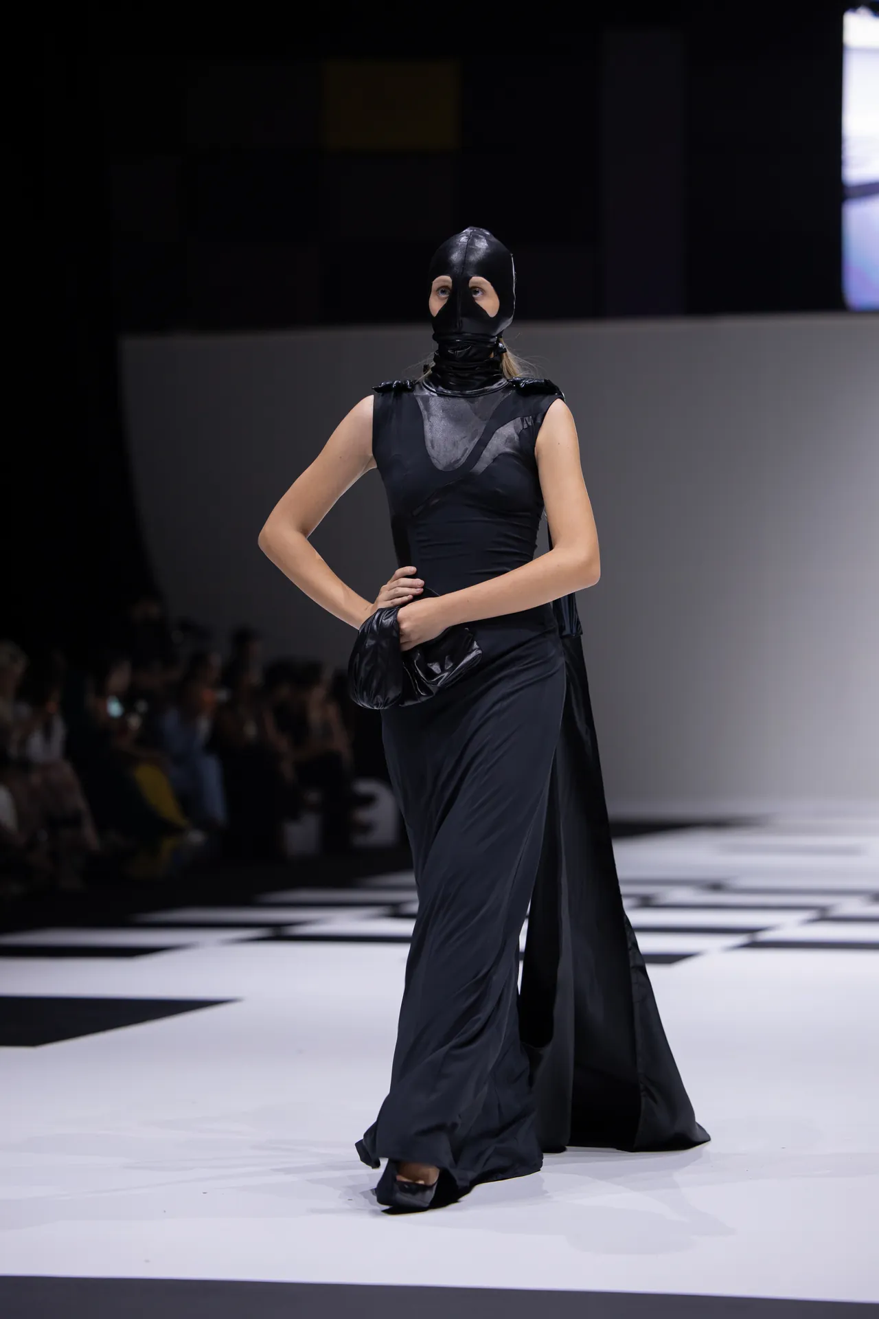 Model walking a runway in an all-black, dramatic gown with a black leather hood/mask and clutch, on a checkered stage.