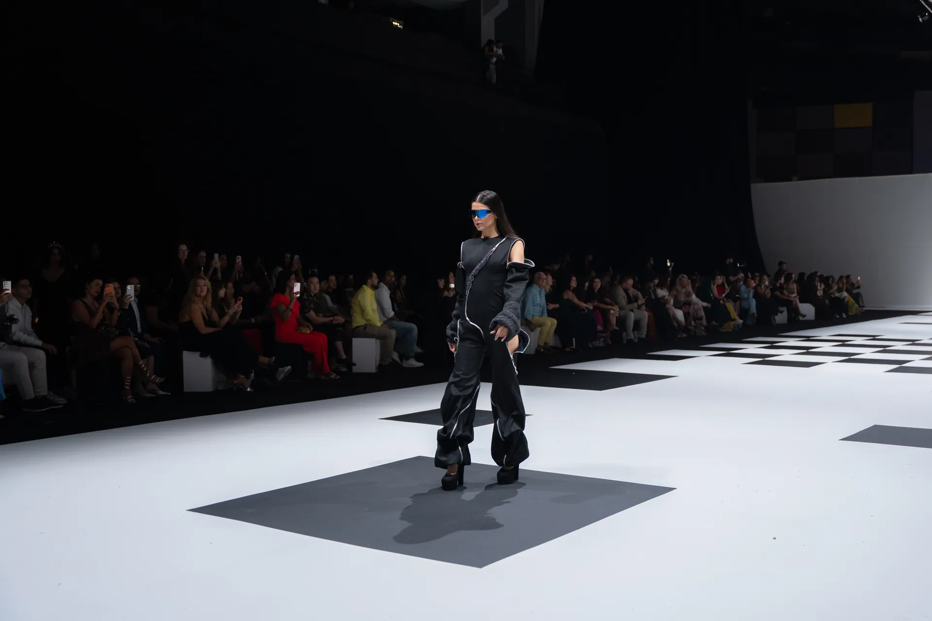 Model walks a fashion runway in a black avant-garde outfit with blue visor sunglasses, as the audience watches from the left.