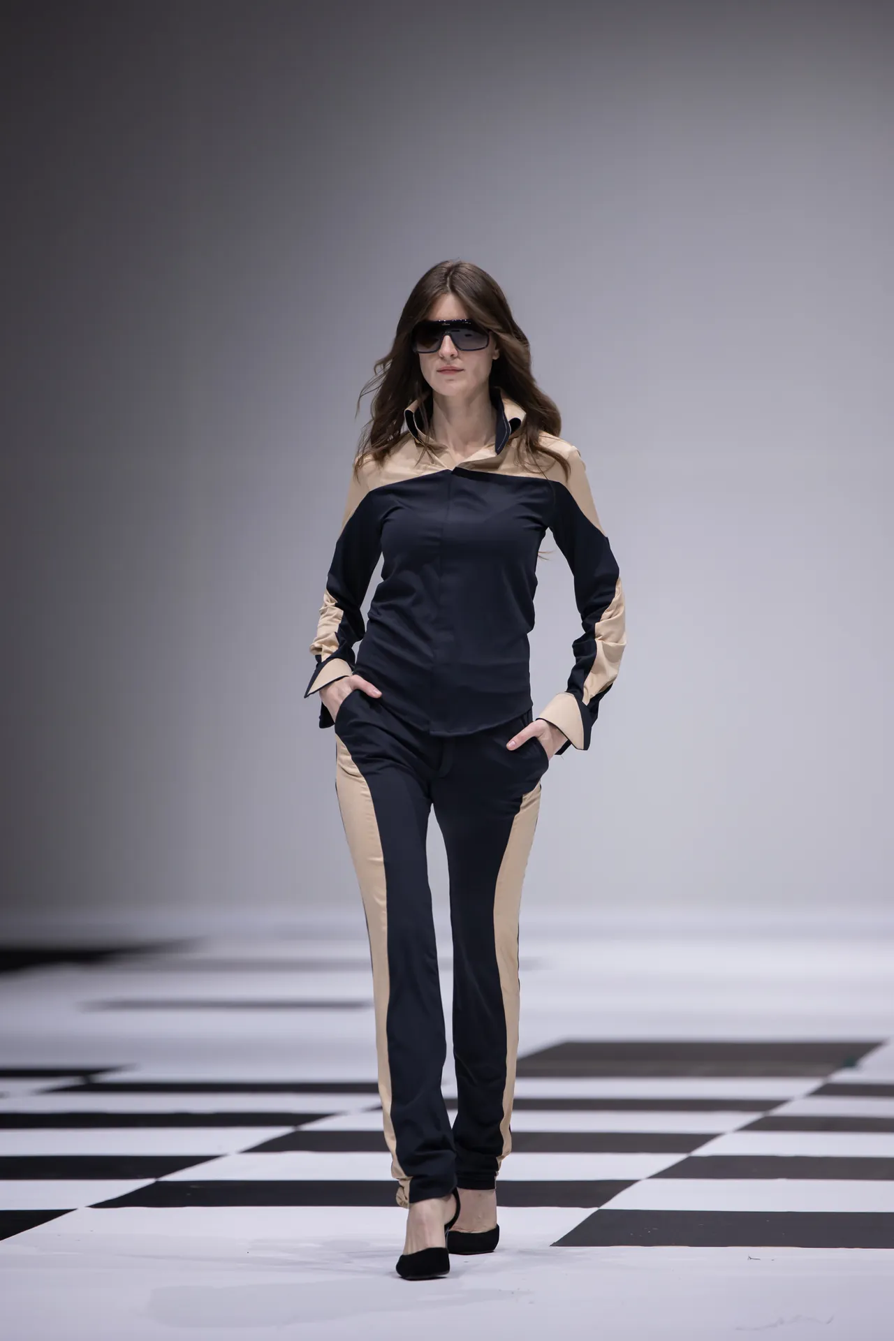 Model walking down a runway in a beige and navy color-block outfit with dark sunglasses, on a black-and-white checkered catwalk.