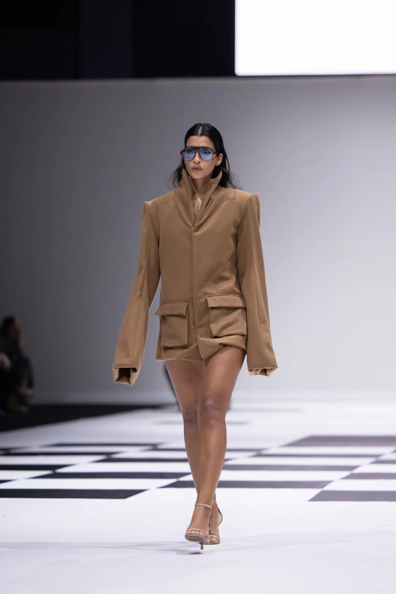 Female model walks the runway wearing a camel oversized blazer with large front pockets and sunglasses on.
