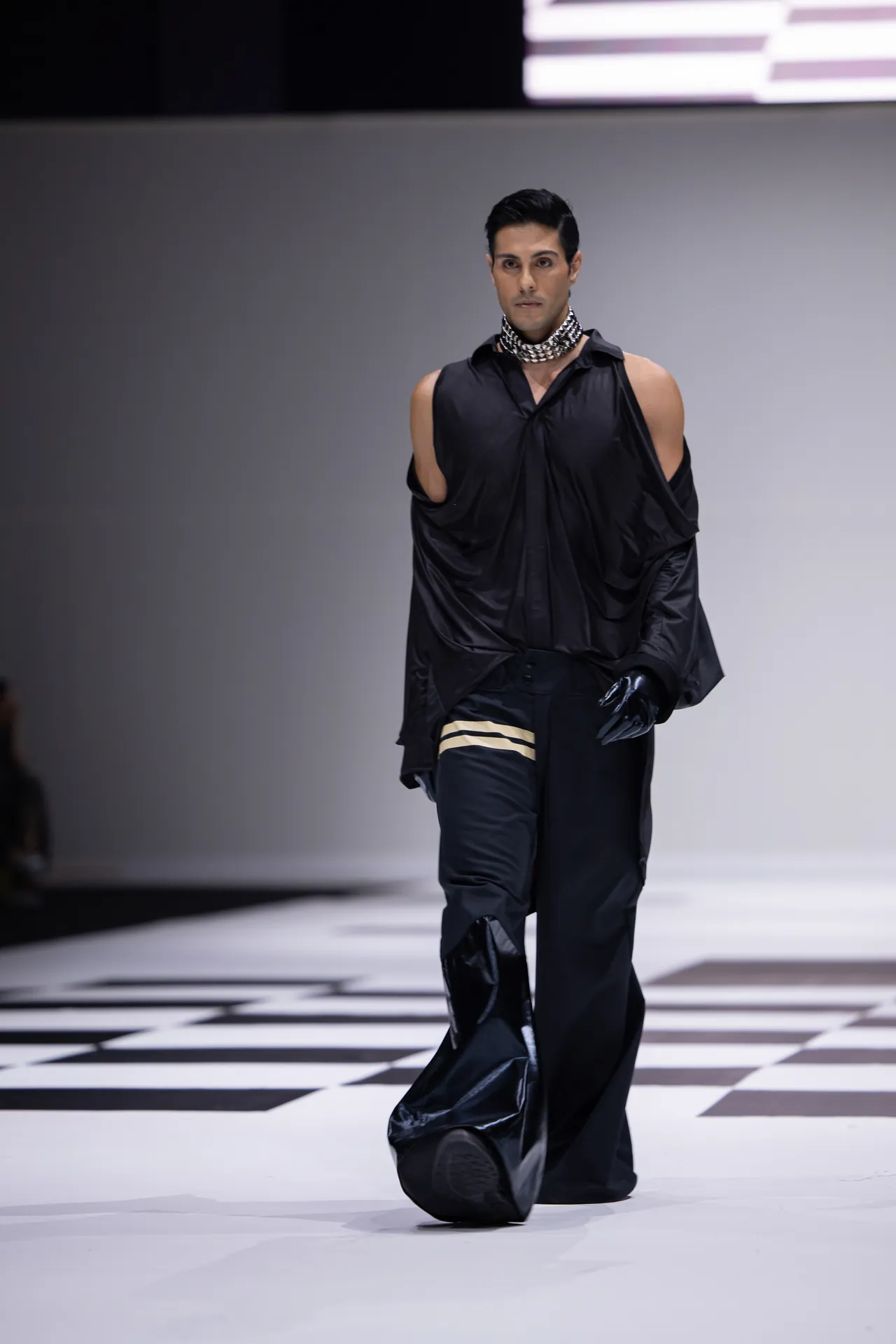 Male model on a black-and-white checkerboard runway wearing an oversized black shirt with exposed shoulders, black pants, gloves, and a chunky chain choker.