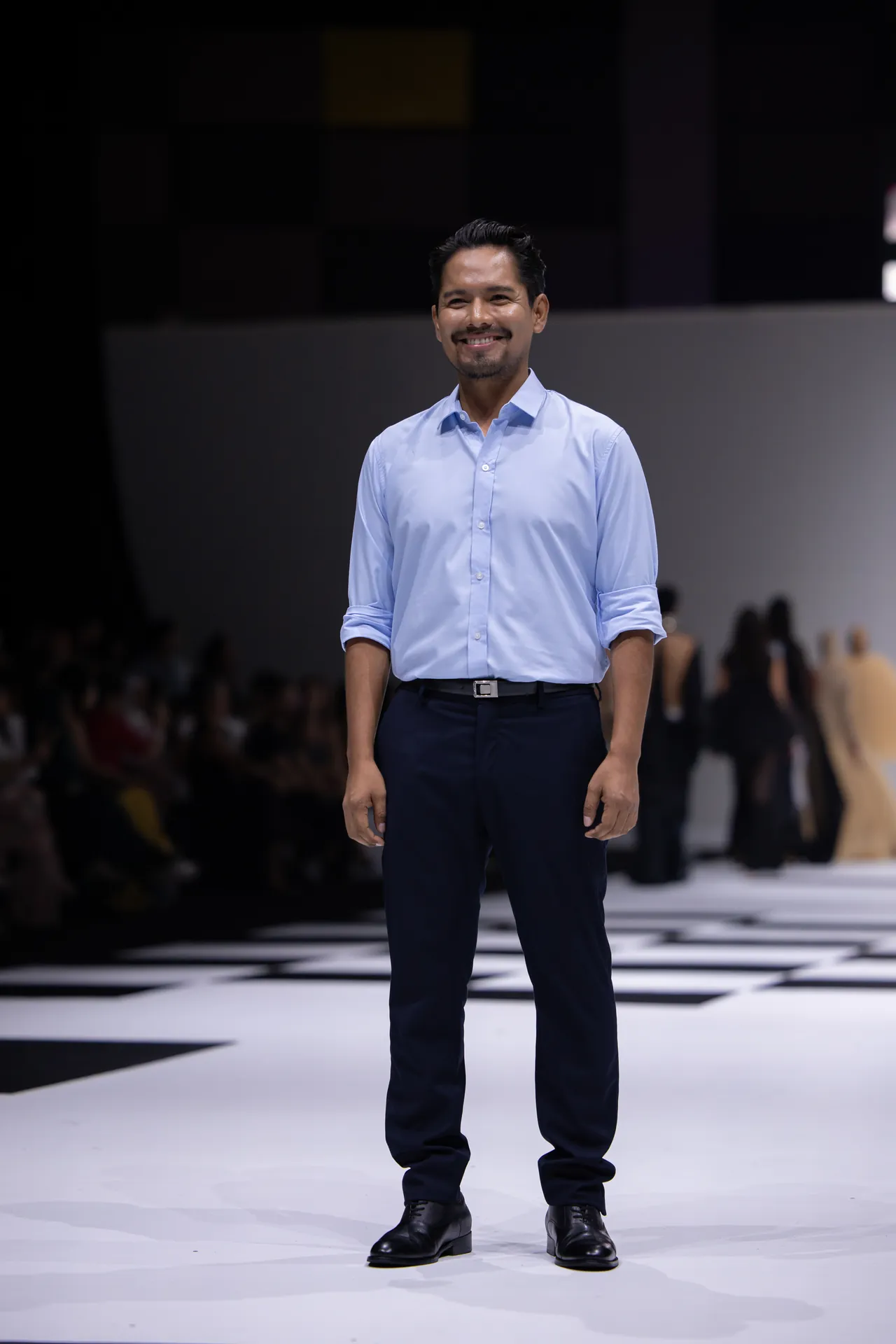 Man in a light blue shirt and dark trousers standing on a fashion runway, smiling, with a checkered floor and blurred audience in the background.
