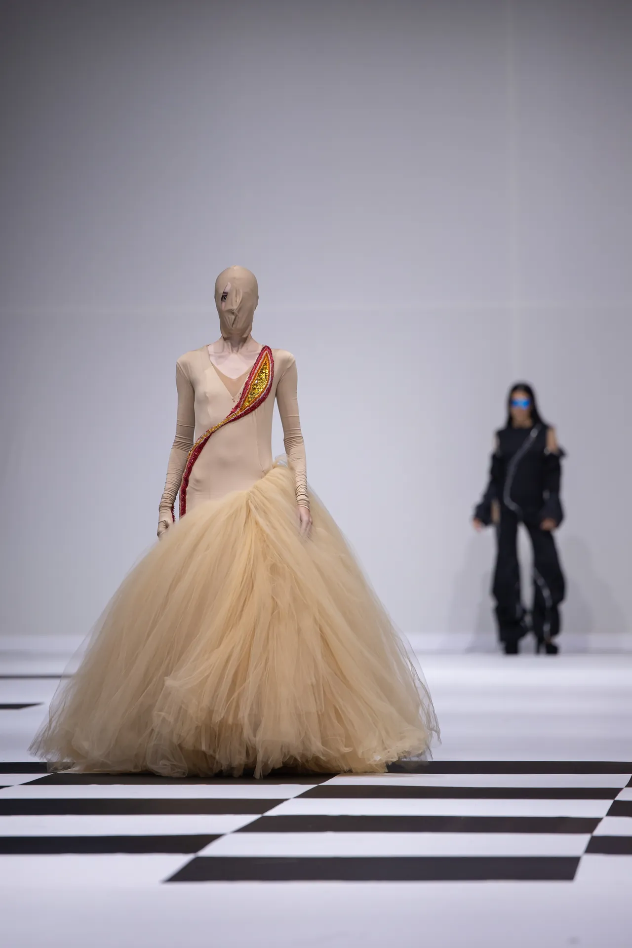 Model walks a runway in a champagne beige gown with a dramatic cream tulle skirt and a red-sequin sash. Another figure in dark clothing stands in the background.