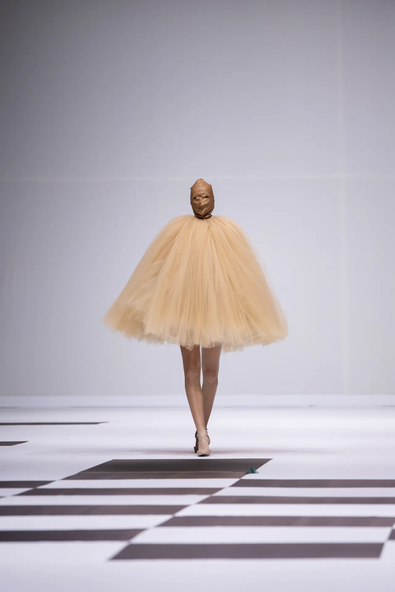 Model walking a fashion runway in a voluminous beige tulle dress with a brown balloon-like headpiece.