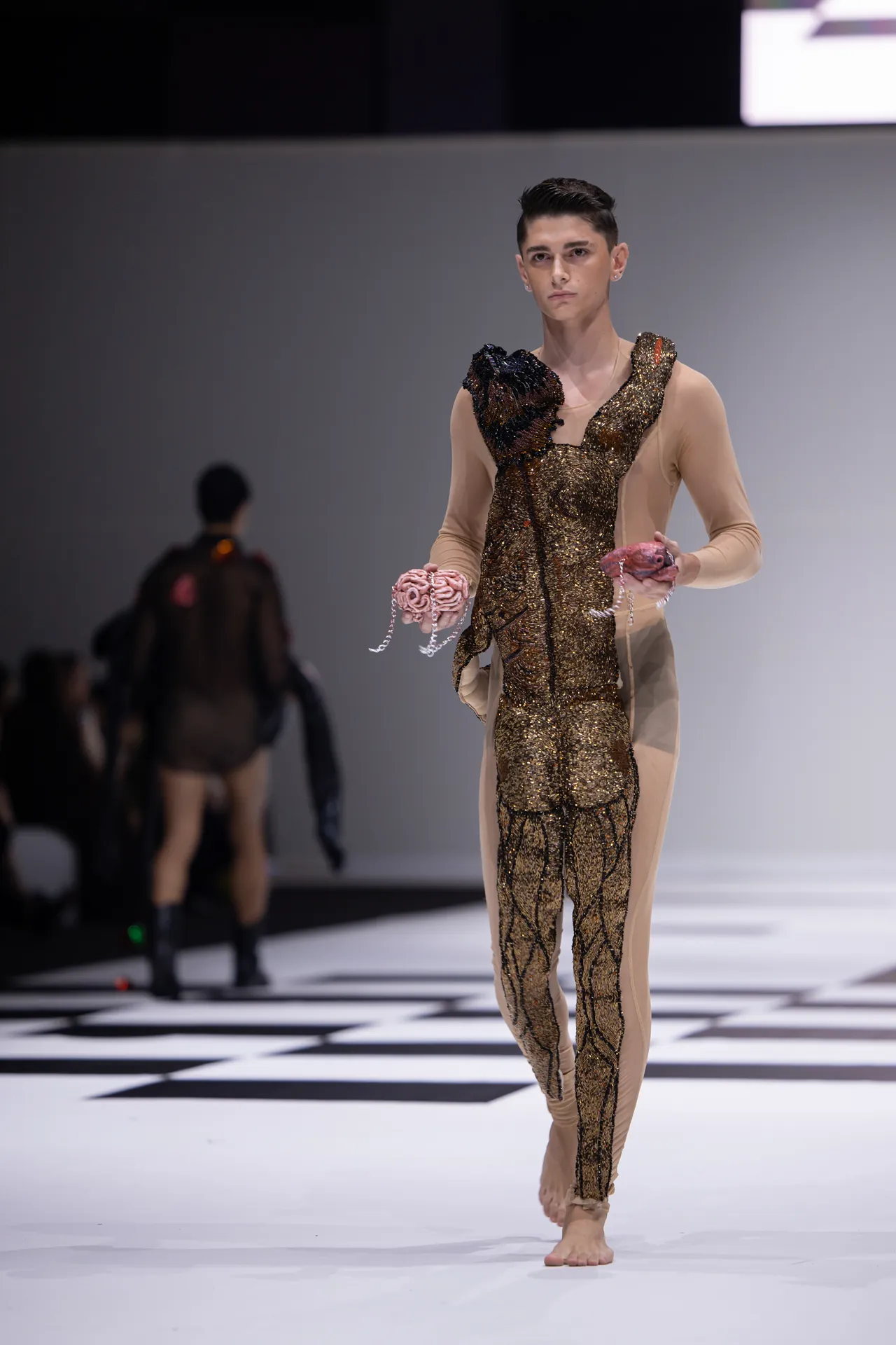 Male runway model in a gold sequin bodysuit with textured black/brown shoulder detail, barefoot on a black-and-white checker stage, holding pink beaded accessories in each hand.