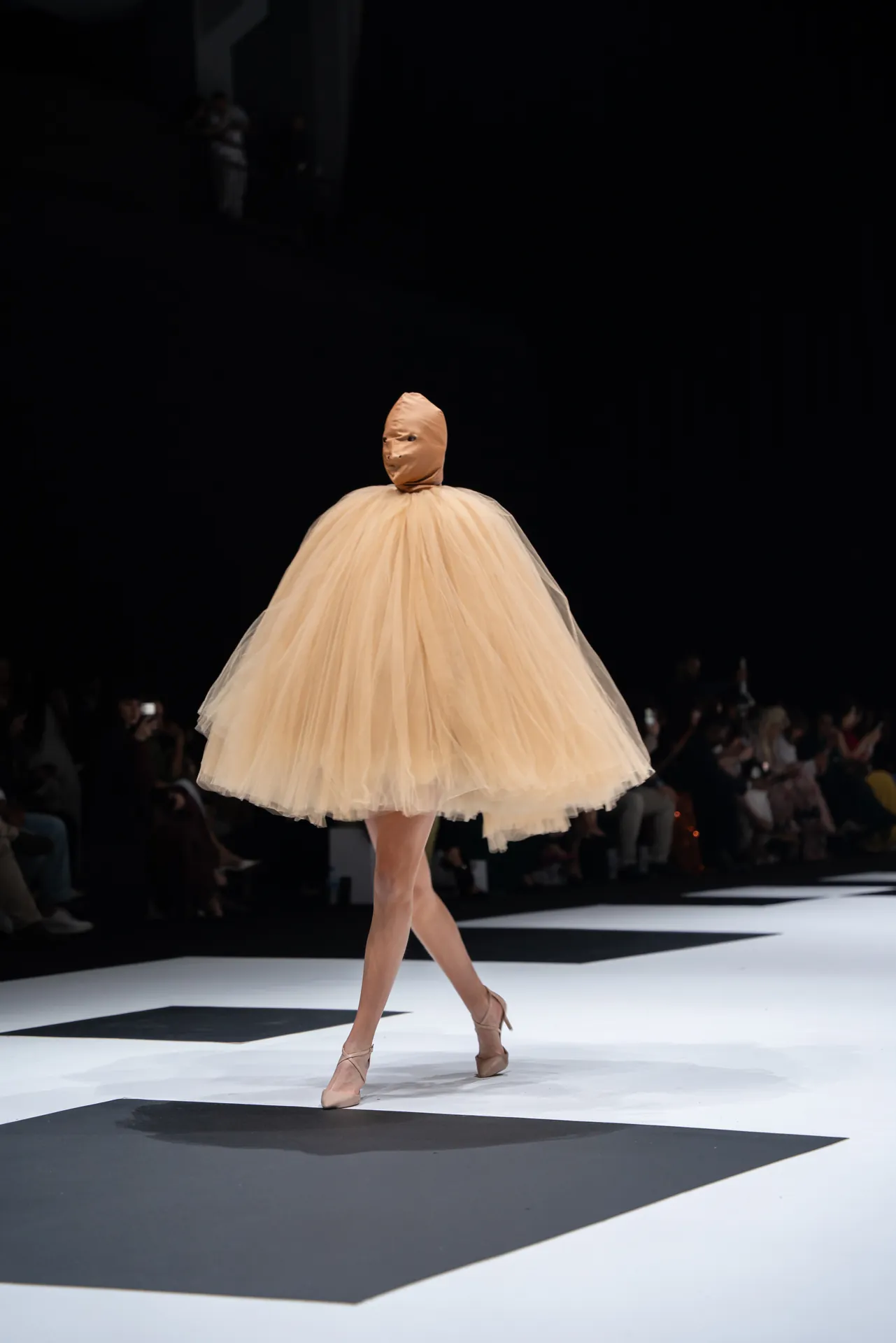 Runway model in a voluminous pale peach tutu and nude headpiece walks the catwalk in beige heels. Audience in dark seating behind.