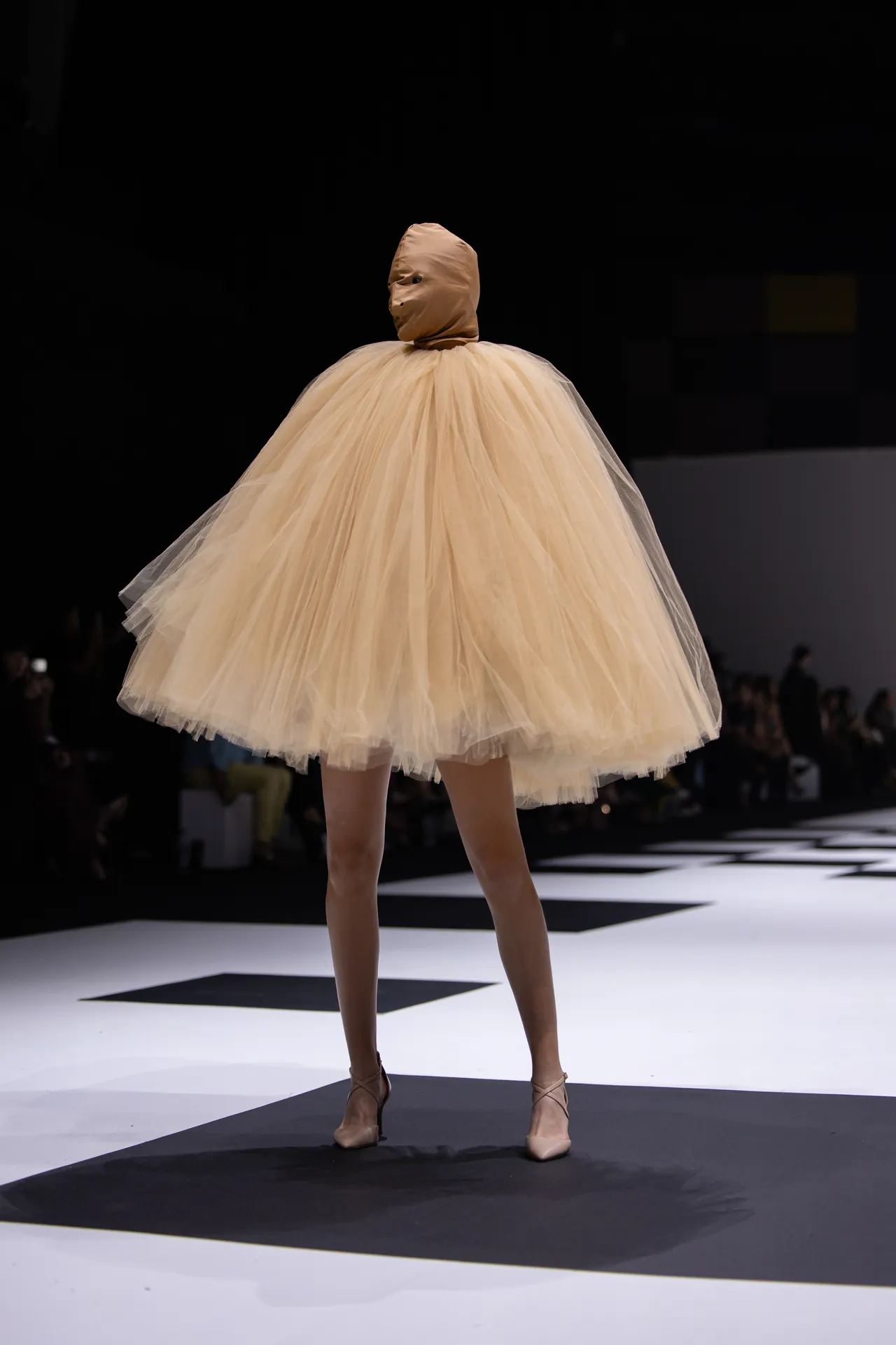 Runway model wearing a pale beige tulle dress with a hooded top, posing on a striped catwalk.