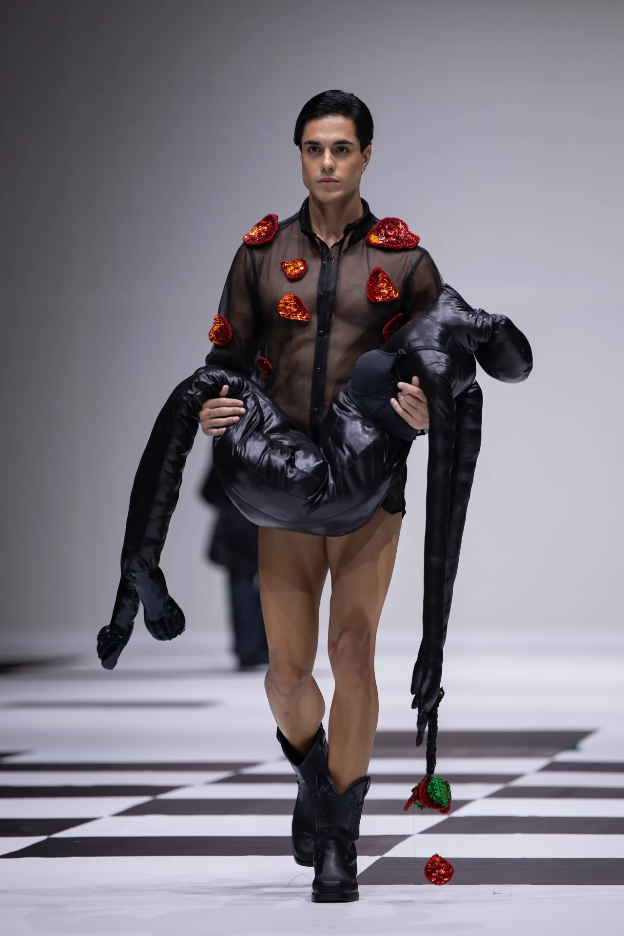Male model on a fashion runway wearing a sheer black shirt with red sequin heart patches, tan tights, and black boots, holding a large black inflatable prop.