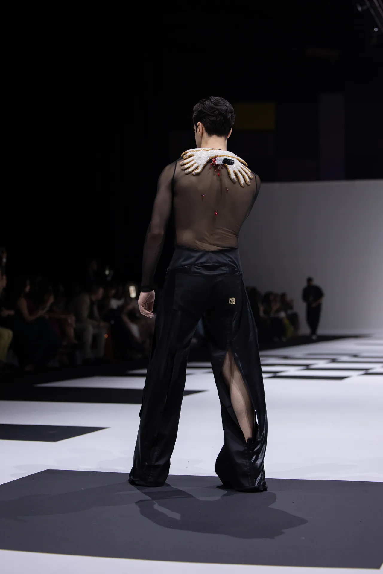 Runway model viewed from behind in a sheer black top and wide black pants, with a white beaded shoulder adornment resting across his shoulders.