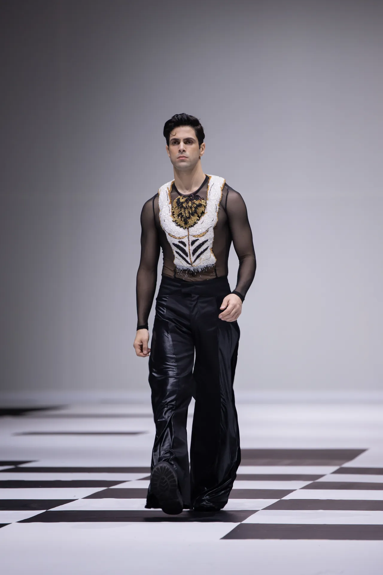 Male fashion model walking the runway in a sheer black mesh top with an ornate white-gold embroidered vest, black pants, on a black-and-white checkerboard floor.