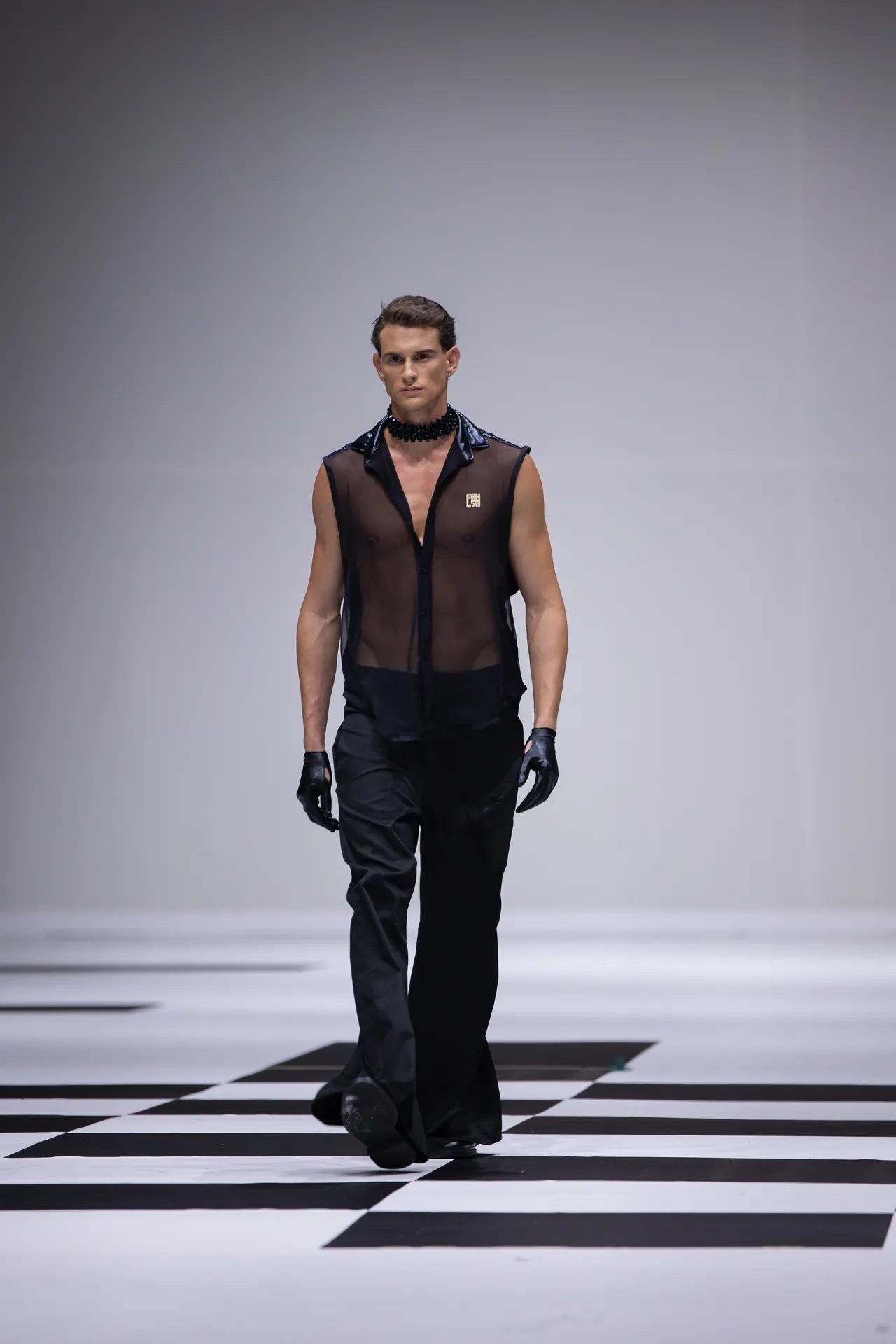 Male model walking a fashion runway in a sheer black sleeveless shirt, black pants, gloves, and a chunky black necklace, against a white, minimalist backdrop.