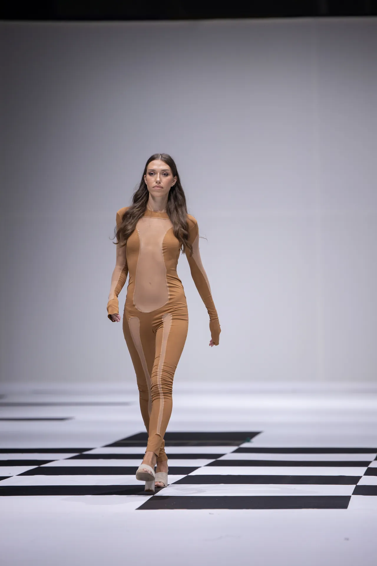 Model walking a beige full-body bodysuit down a black-and-white checkerboard runway.