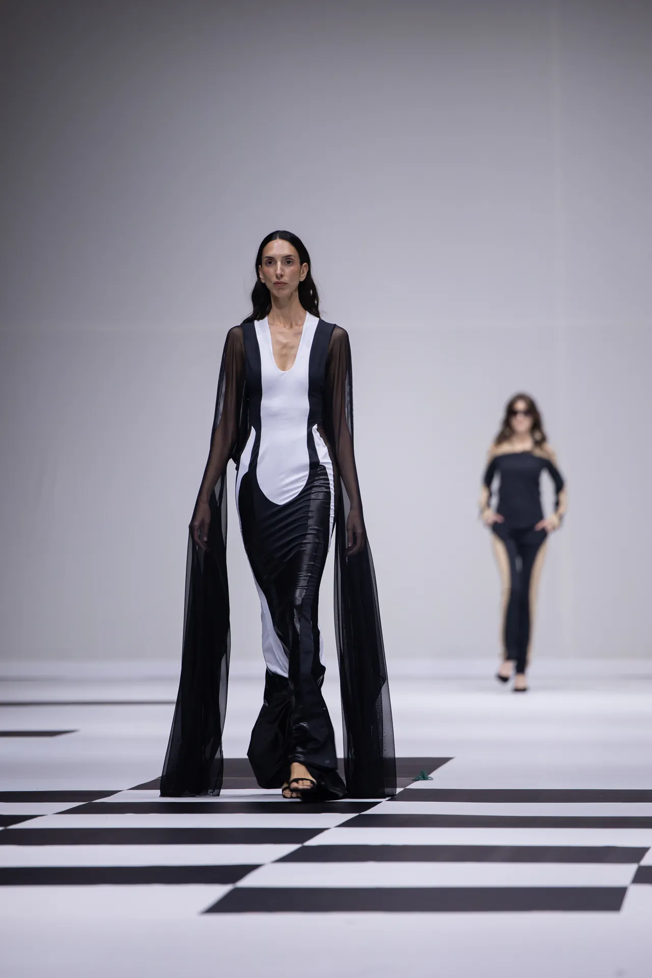 Model walks a black-and-white evening gown with a sheer cape on a checkered runway in a fashion show.