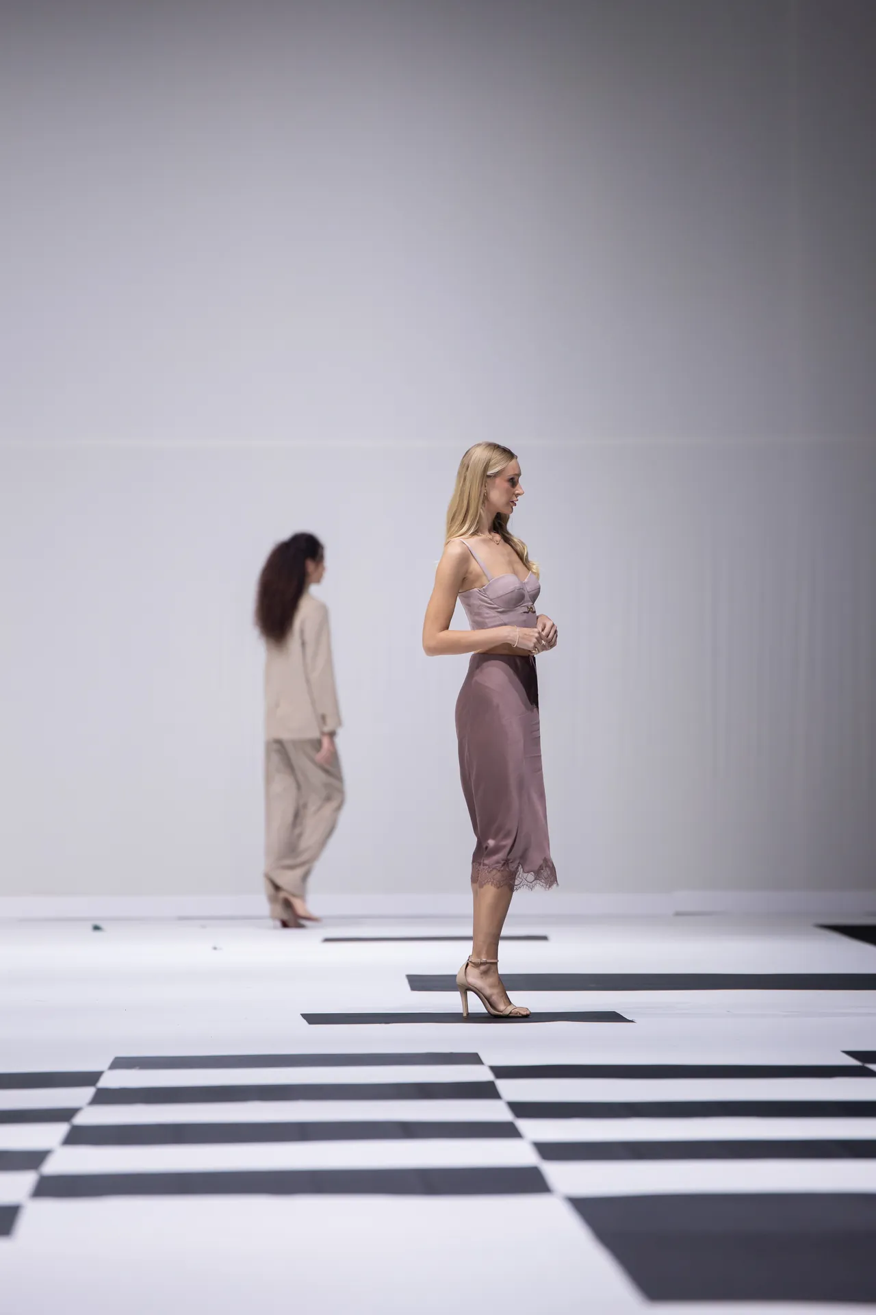 Female model in a pink satin dress and high heels walks on a black-and-white checkerboard runway; another model blurred in the background.