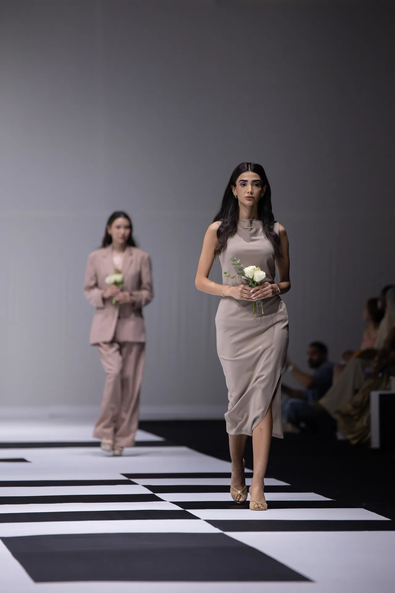 Model in a beige satin dress struts a black-and-white runway, holding a small bouquet of white roses; blurred second model behind.