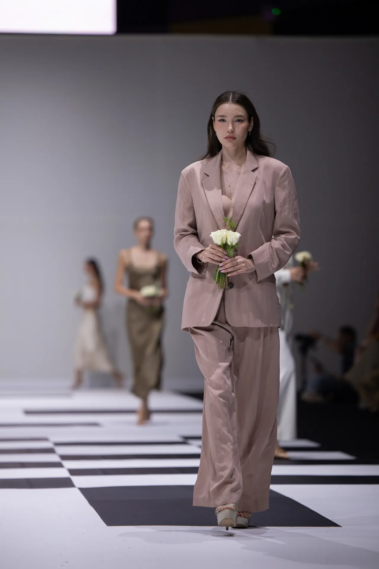 Model in a blush pink suit walks the runway, holding a single white rose, with blurred background models.