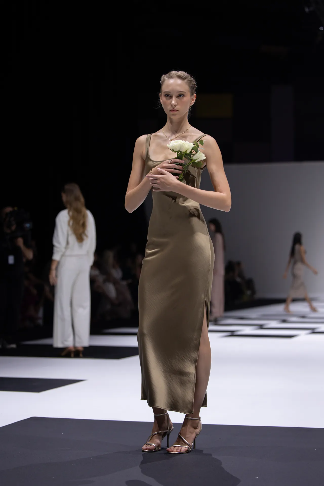 Model on a runway wearing a gold satin slip dress and strappy sandals, holding white roses under bright stage lights.