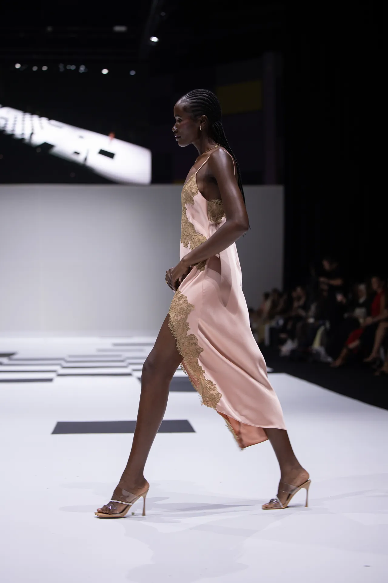 Model walking a fashion show runway in a pale pink satin dress with gold lace trim and clear heeled sandals, side profile view.