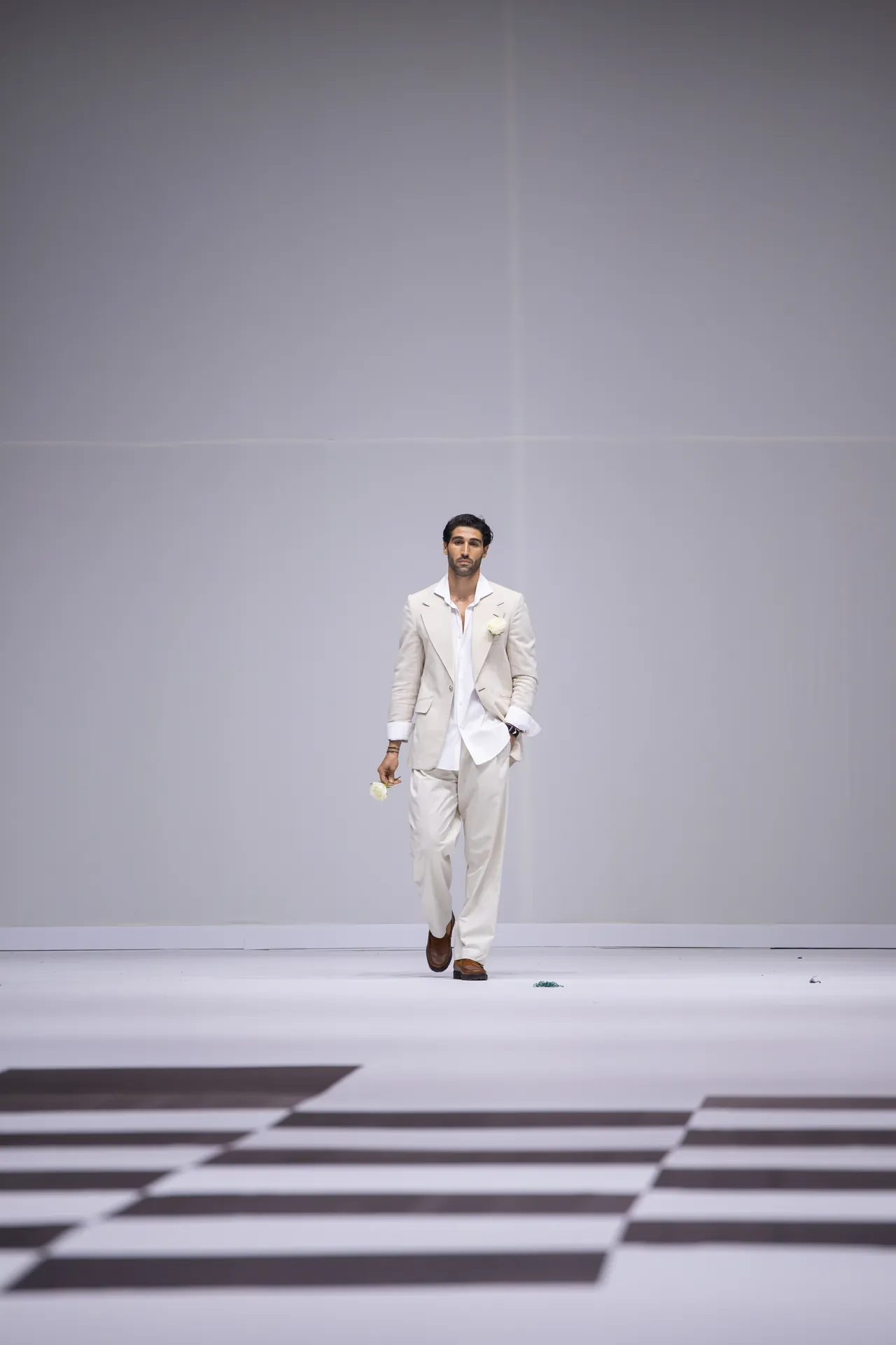 Male model in a light beige suit walking down a fashion runway, holding a white flower in each hand against a minimalist backdrop.