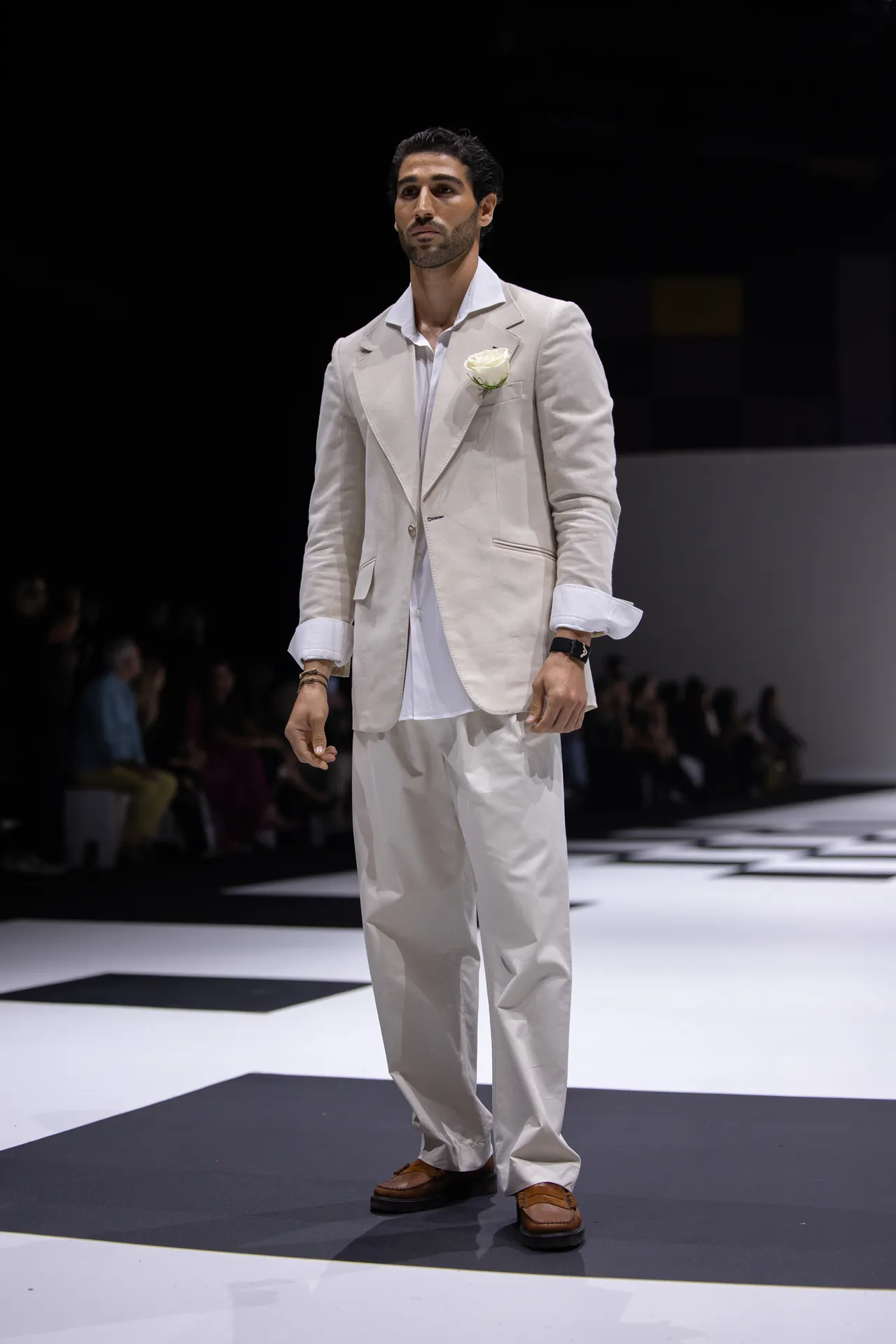 Male model walking a fashion runway in a cream suit with white shirt and boutonniere, brown loafers visible against a black-and-white runway backdrop
