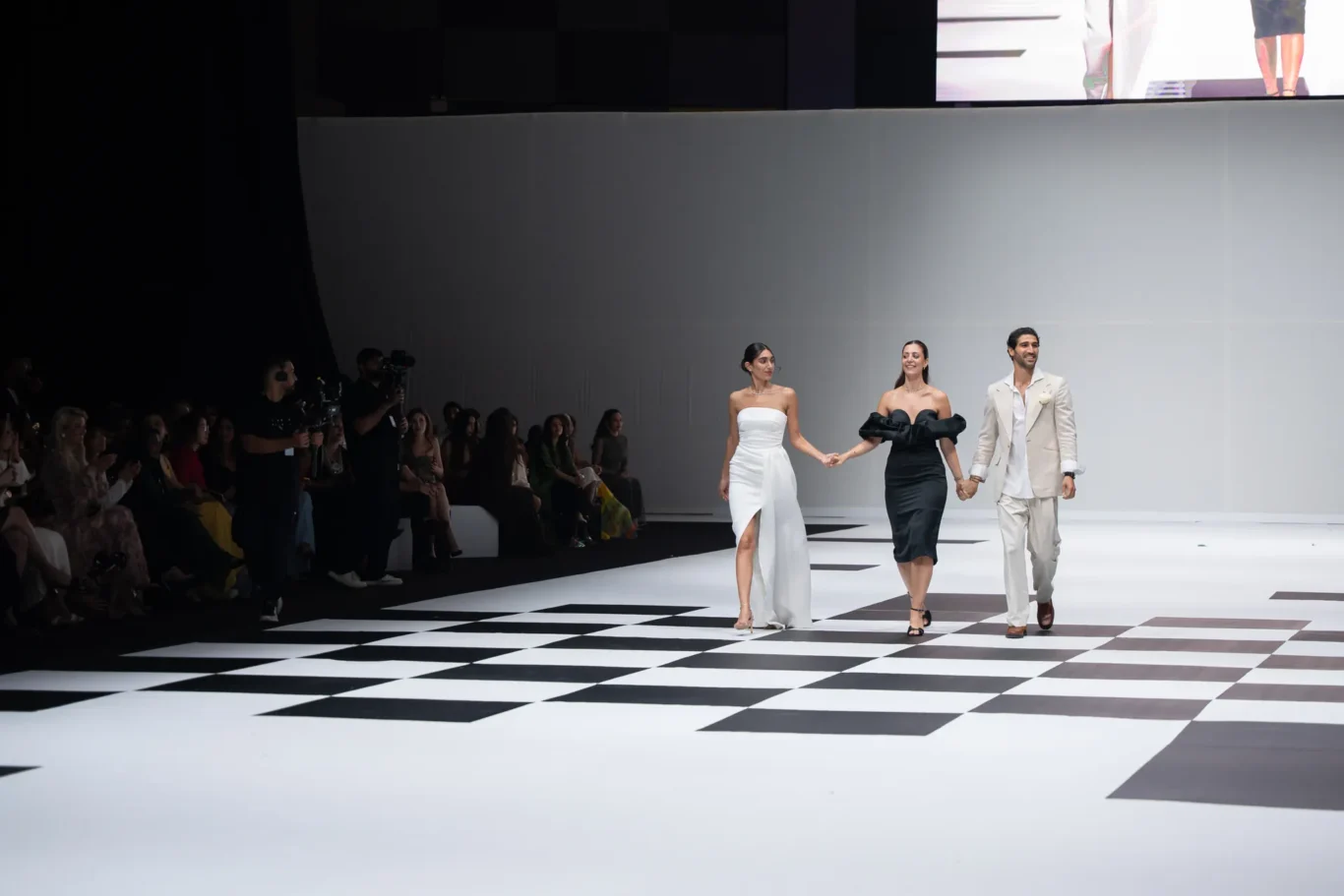 Three models and a man walk hand in hand down a black-and-white checkered runway as a seated audience watches from the left.