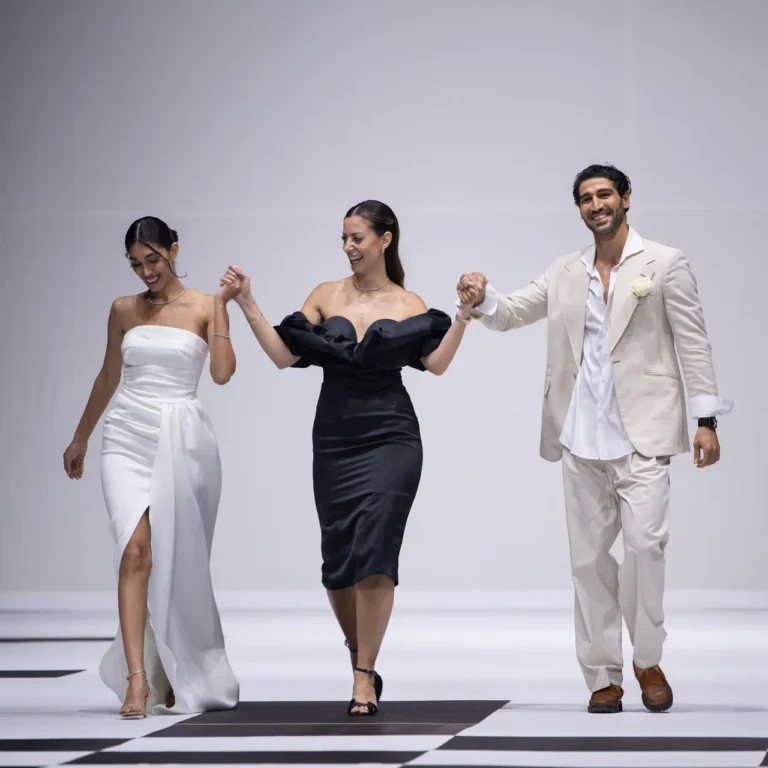 Three models walk hand-in-hand on a black-and-white checkerboard runway; woman in a white strapless gown, woman in a black off-shoulder dress, man in a cream suit.