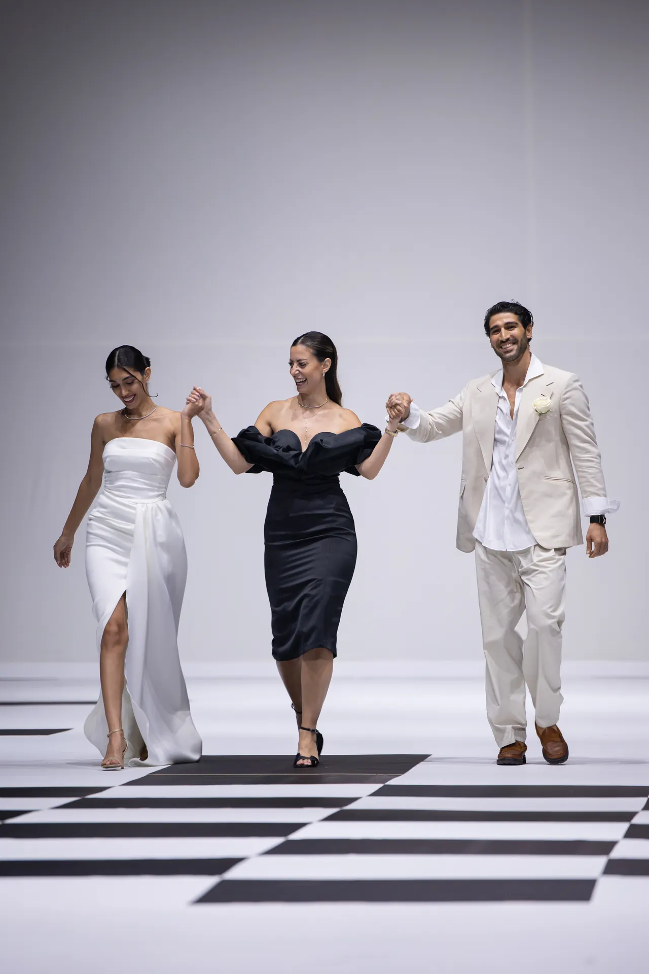 Three models walk hand-in-hand on a black-and-white checkerboard runway; woman in a white strapless gown, woman in a black off-shoulder dress, man in a cream suit.