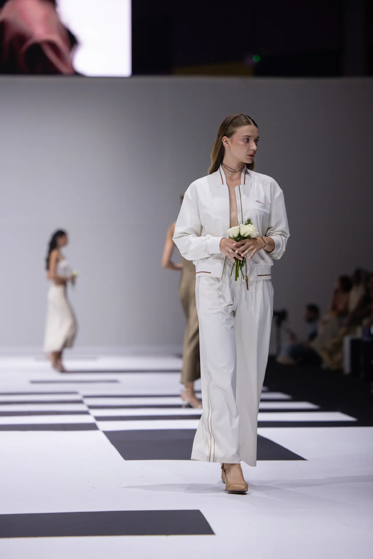 Model walking a fashion runway in a white jumpsuit, holding a small bouquet of white roses under bright lights.
