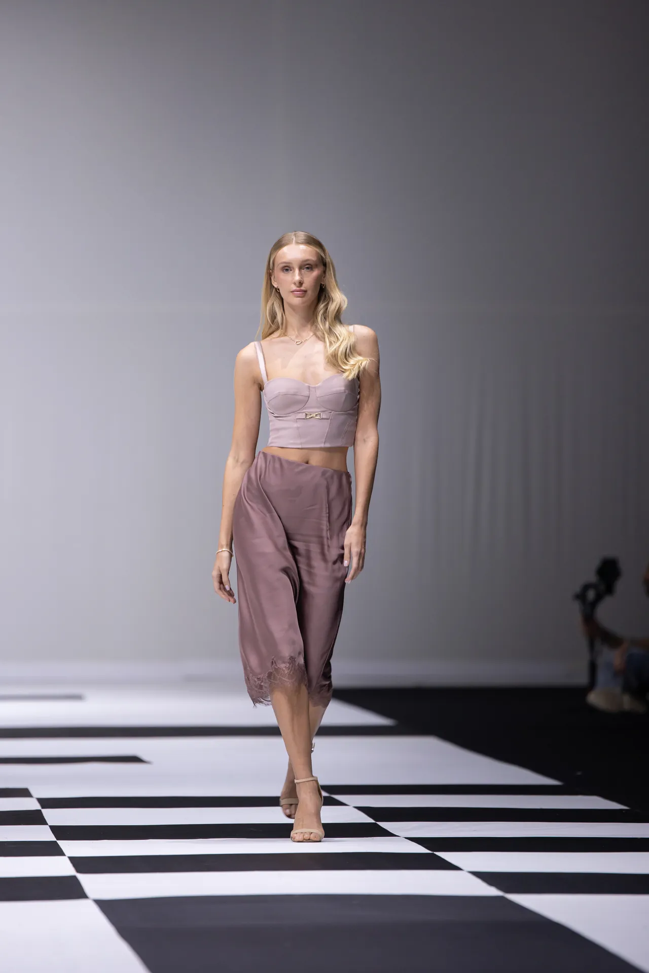 Model walks down a black-and-white runway in a pale pink crop top and satin pants with lace hem.