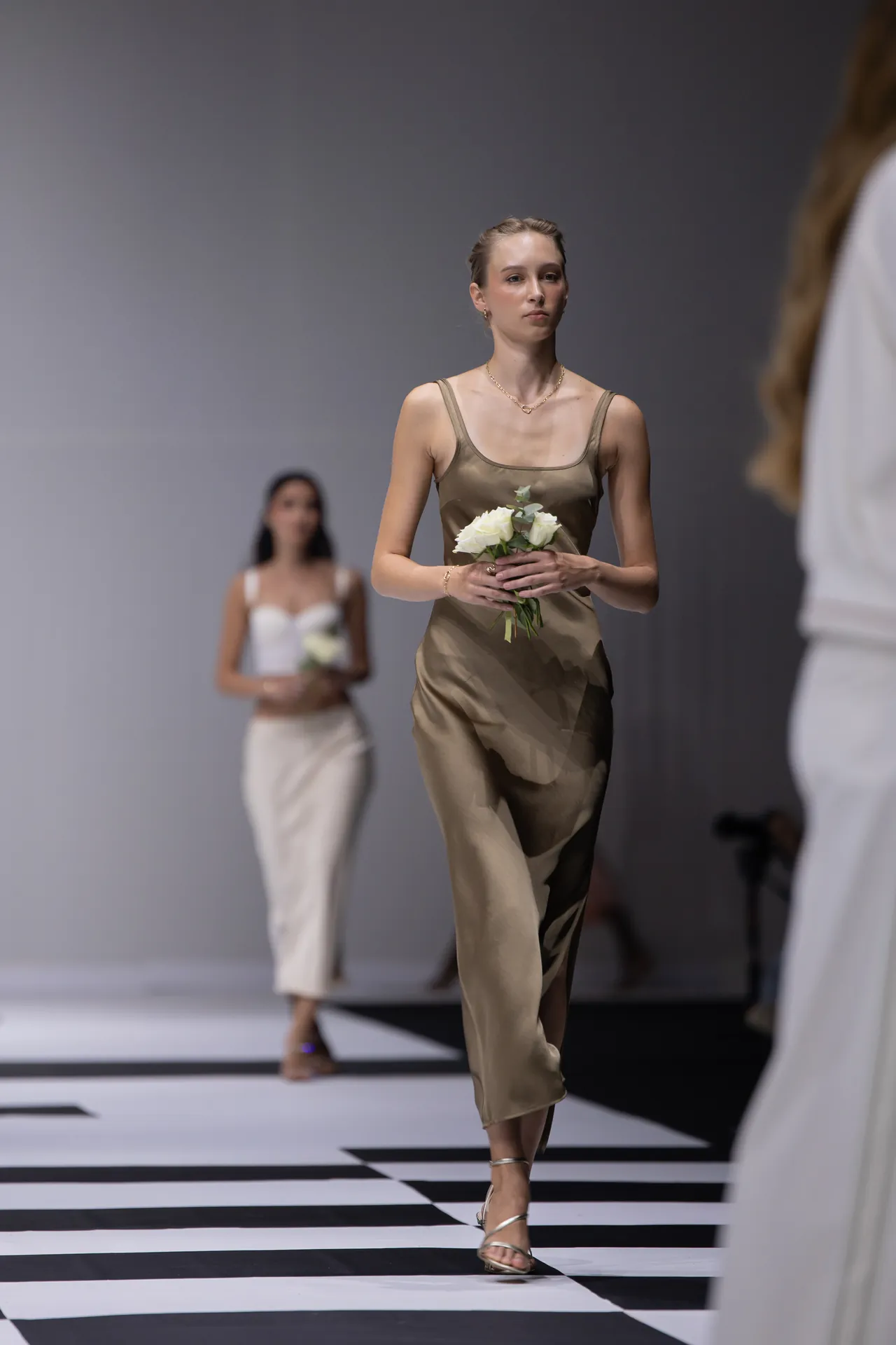 Model in a bronze satin slip dress walks a black-and-white checkered runway, holding a small white bouquet.