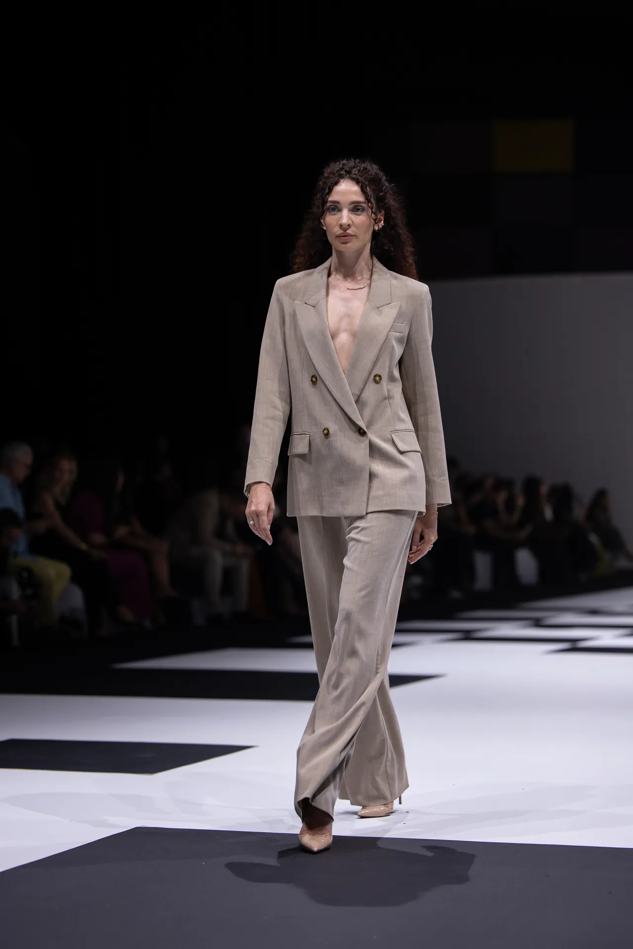 Female model walks a runway wearing a light beige double-breasted pantsuit with wide lapels.