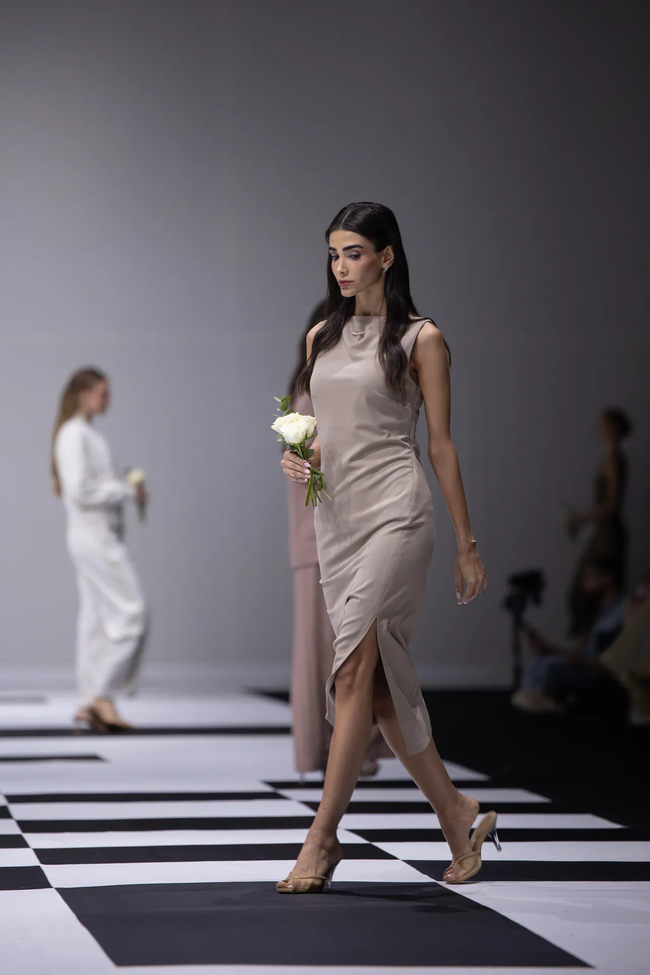 Model walks a taupe satin dress down a black-and-white striped runway, holding a single white rose-focused forward pose.