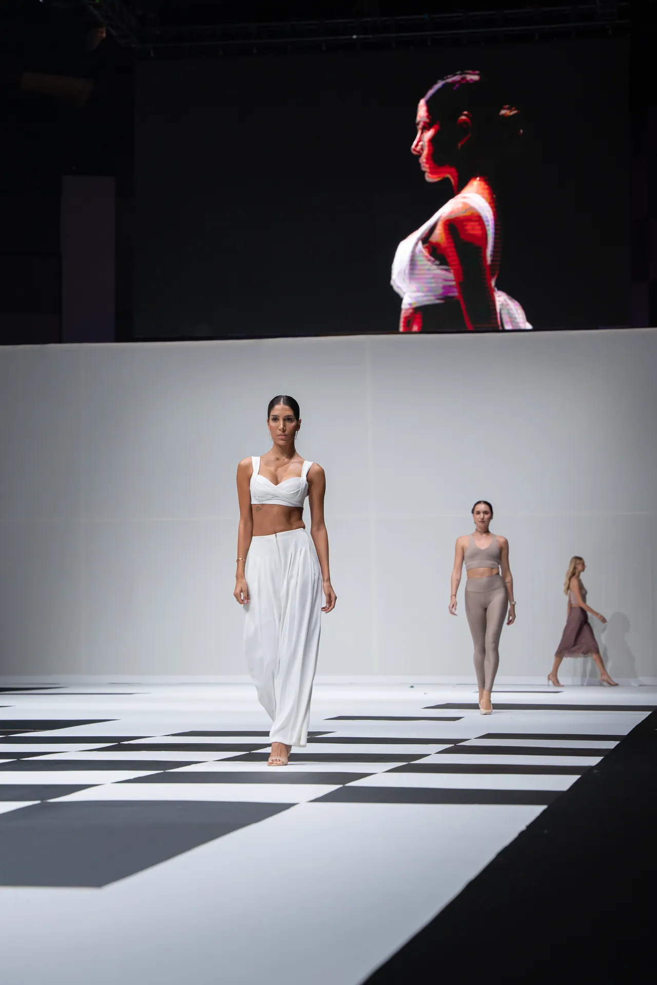 Model walks the runway in a white two-piece outfit as others follow on a black-and-white checkered stage, with a large screen behind showing her profile.