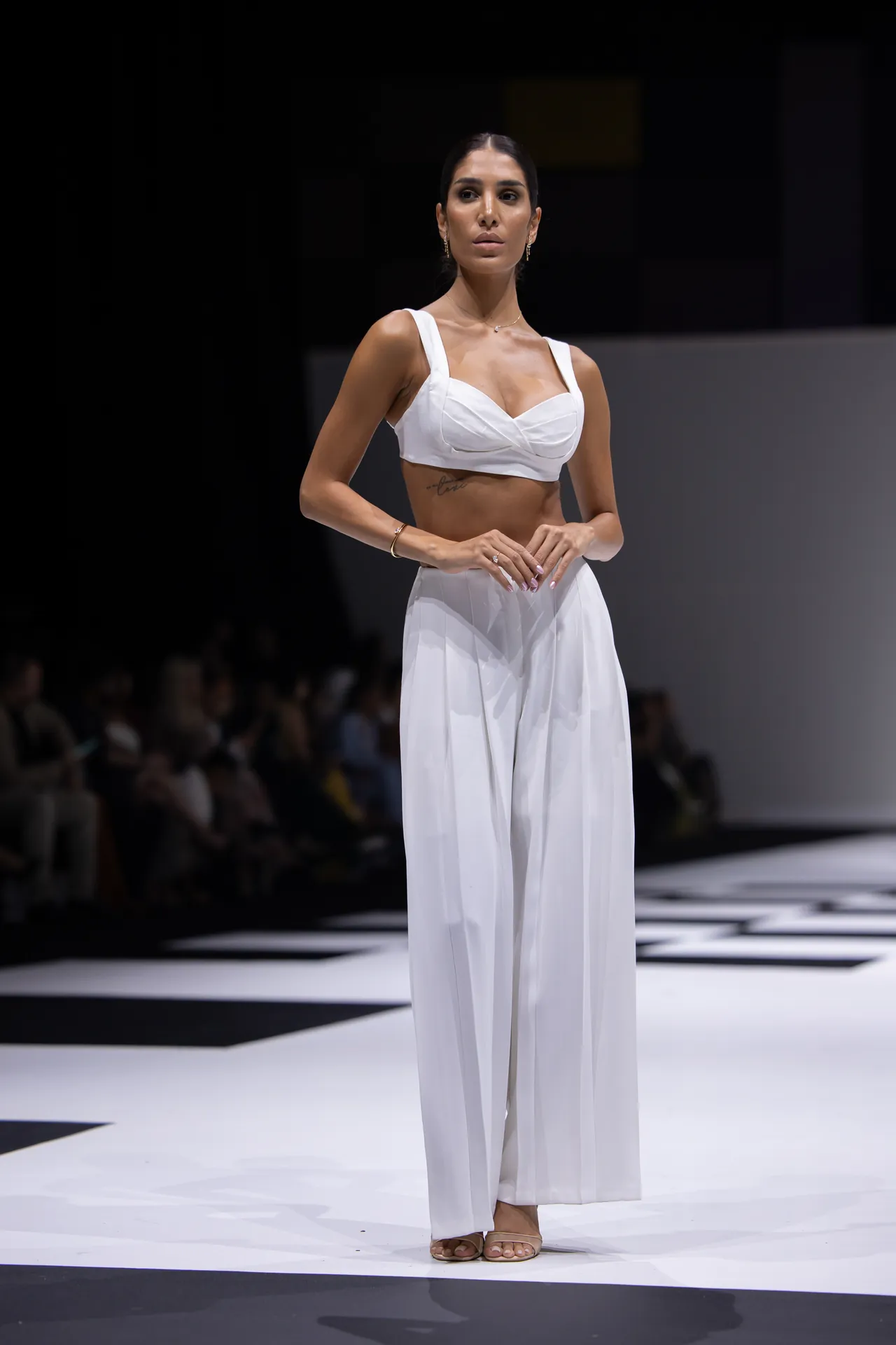 Fashion model on a runway wearing a white crop top and wide-leg pants, hands clasped, spotlight on her.