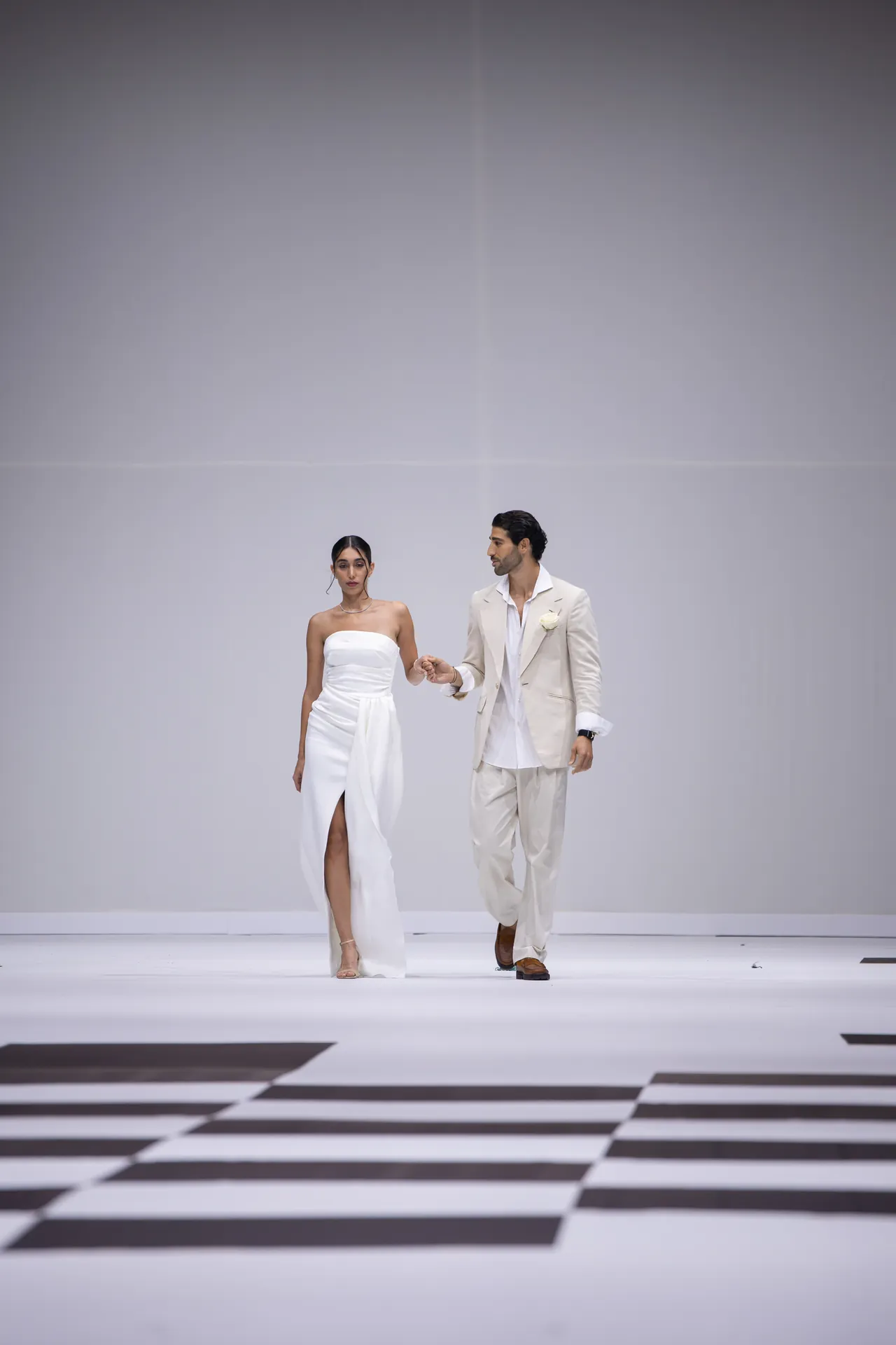 A couple walks on a white runway at a fashion show; woman in a strapless white dress and the man in a light beige suit.