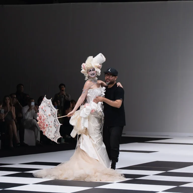 Model in an elaborate white gown with a lace parasol on a fashion runway, escorted by a designer.