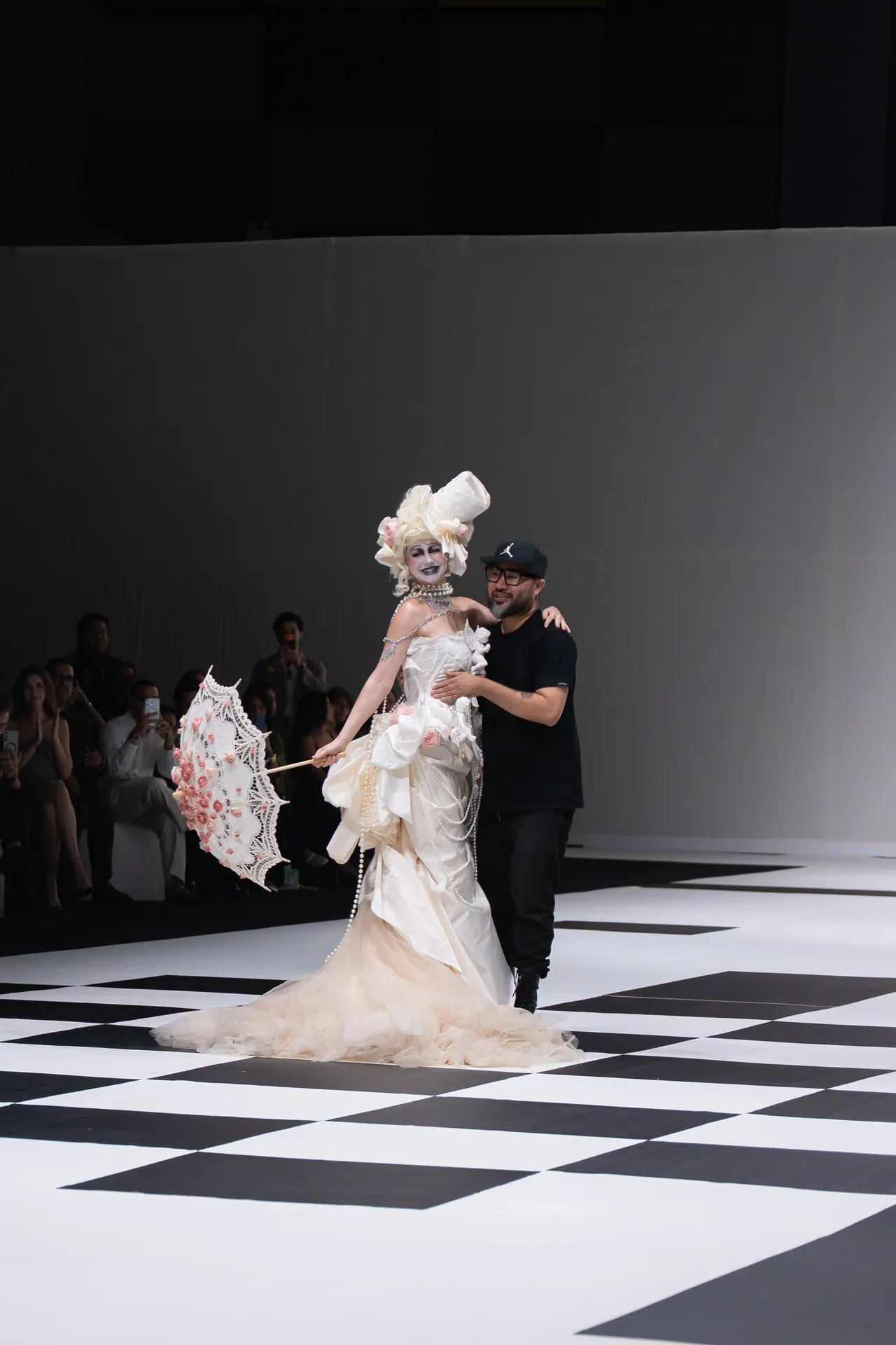 Model in an elaborate white gown with a lace parasol on a fashion runway, escorted by a designer.