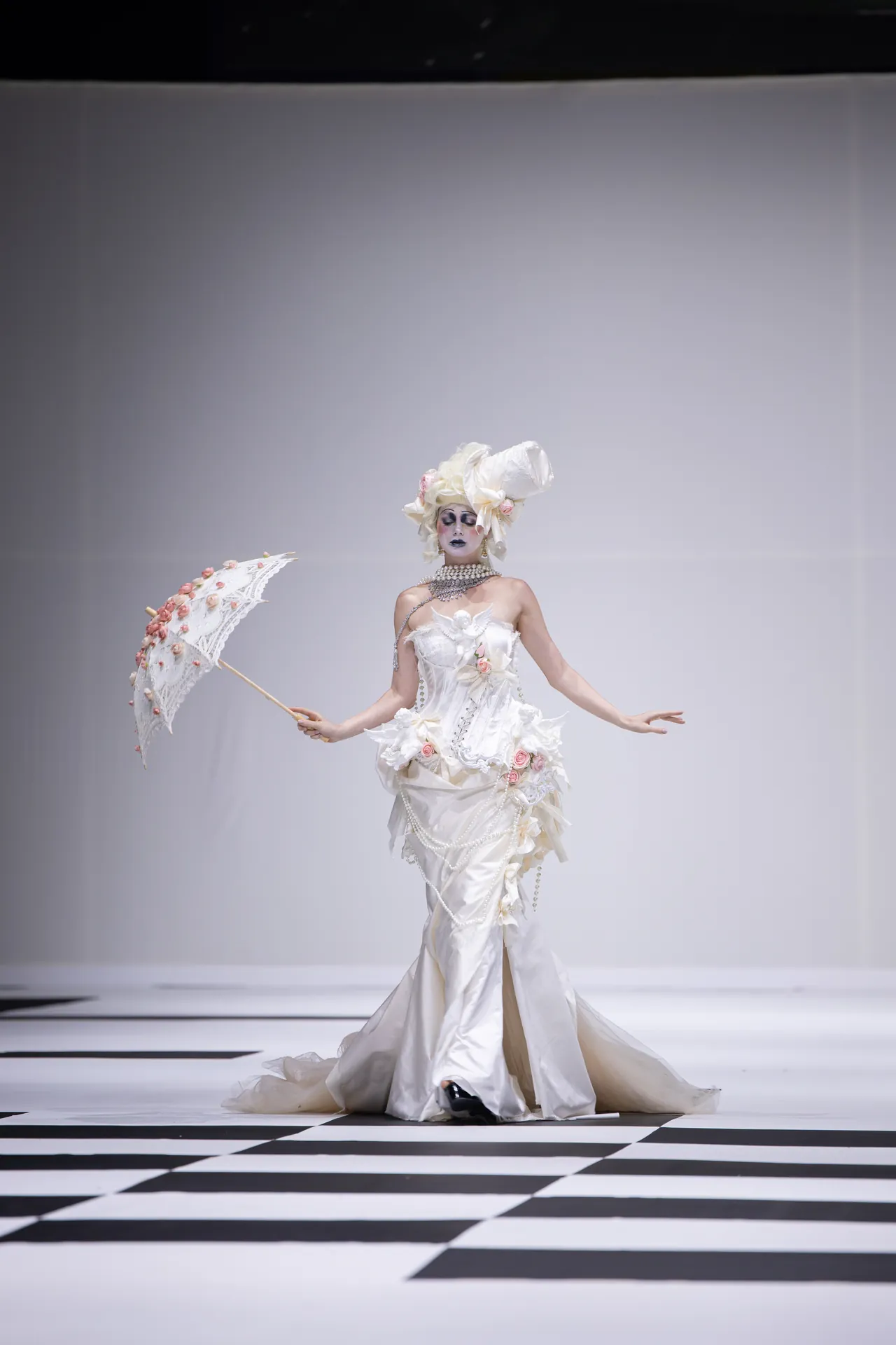 Model in an ornate white couture gown with a large floral headpiece and pearl necklace, holding a lace parasol on a checkered runway.