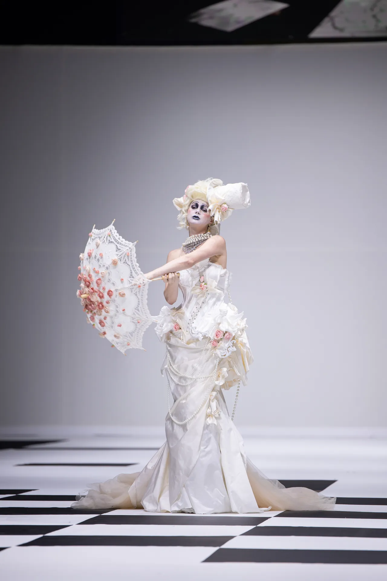 Runway model in an elaborate white gown with a large floral headpiece, pearls, and a lace parasol on a black-and-white checkerboard catwalk.